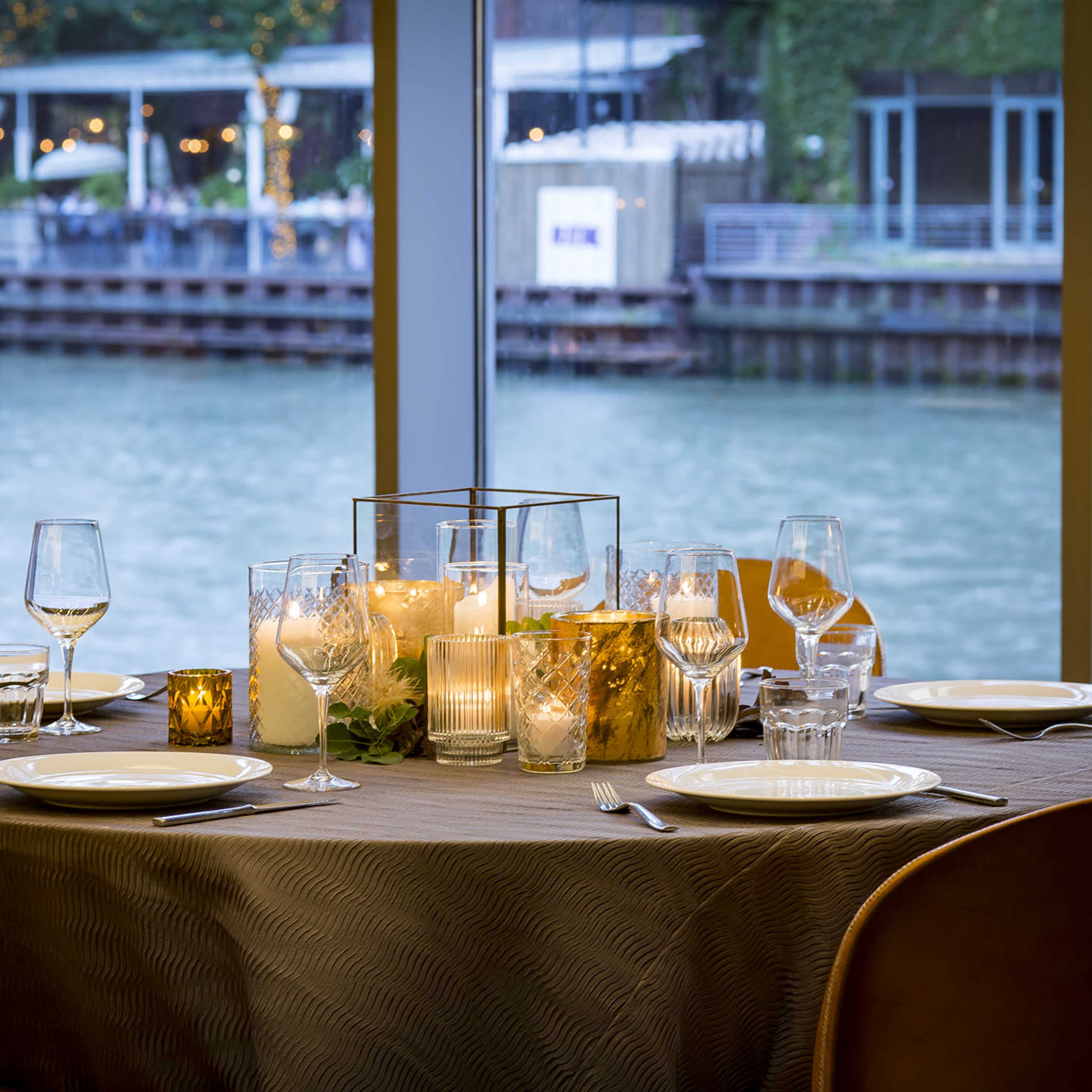 A round dining table is elegantly set with plates, glassware, and candles, overlooking a scenic waterfront through large windows.