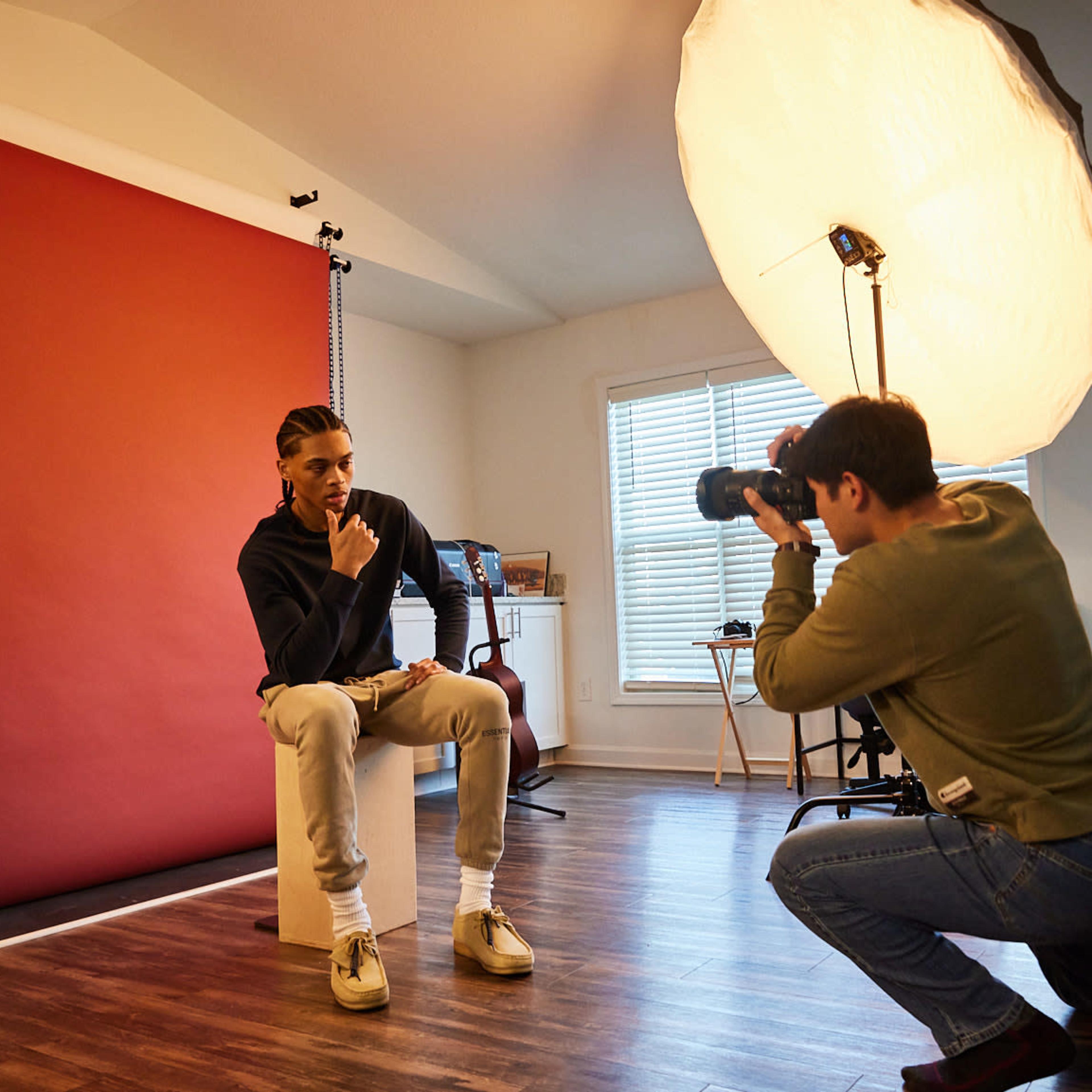 A photographer captures a portrait of a subject sitting on a stool in a studio with a red backdrop and large softbox lighting.