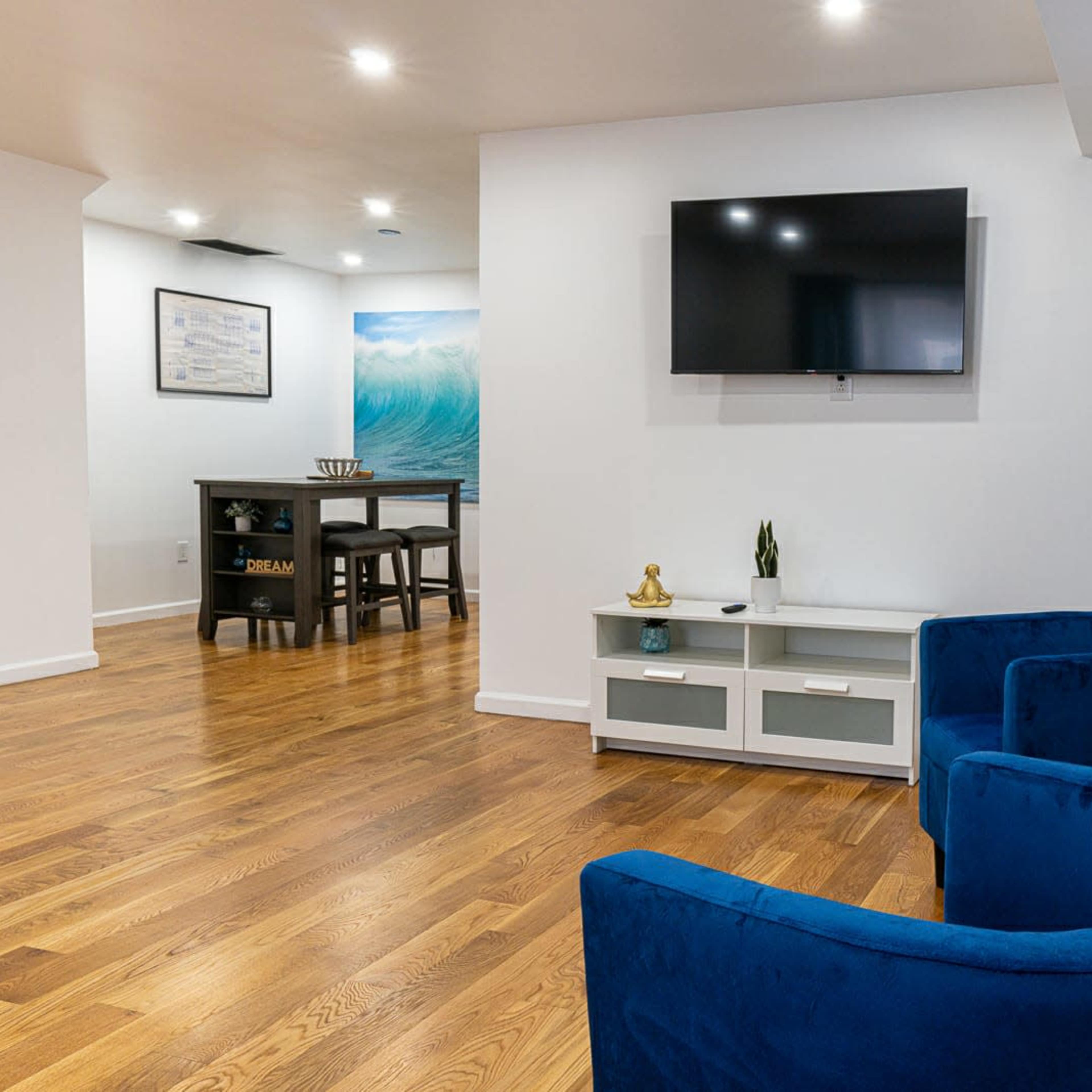 A living space featuring two blue chairs, a white TV unit, and a wall-mounted television, with a dining area visible in the background.