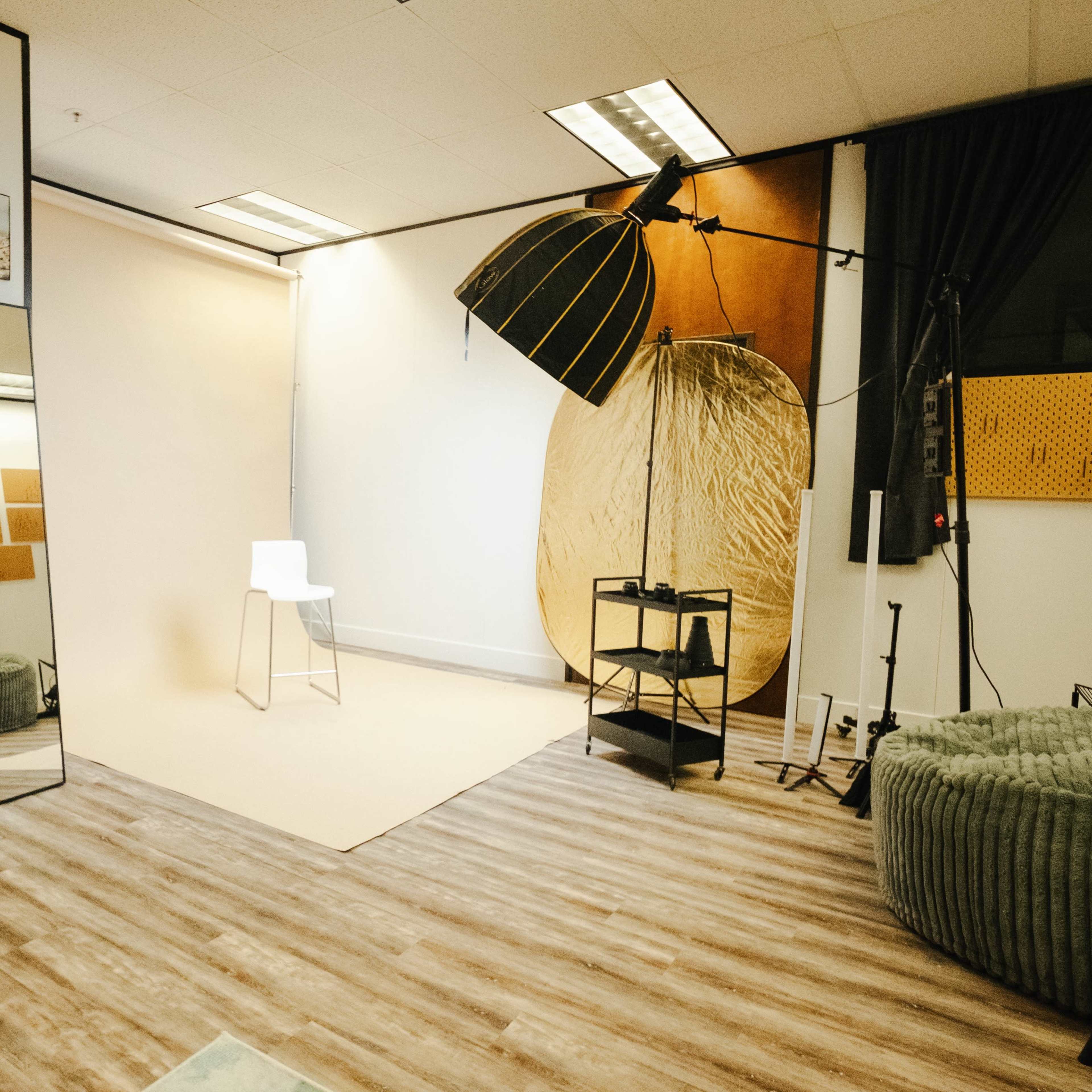 A photography studio equipped with a large mirror, a backdrop, a chair, a stand with lighting equipment, and a textured wall.