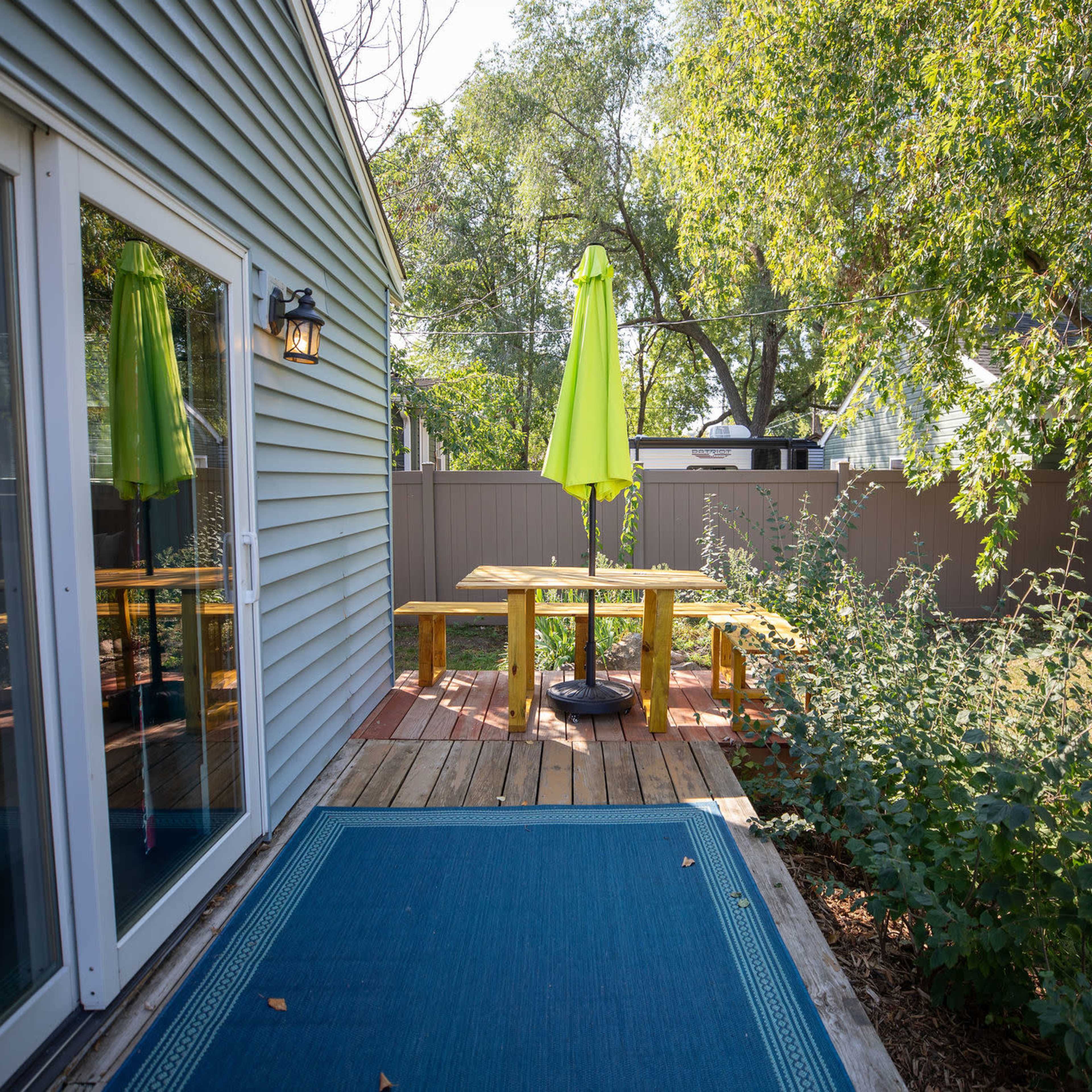 A backyard deck with a yellow umbrella over a wooden table, surrounded by greenery and a light blue rug at the entrance.