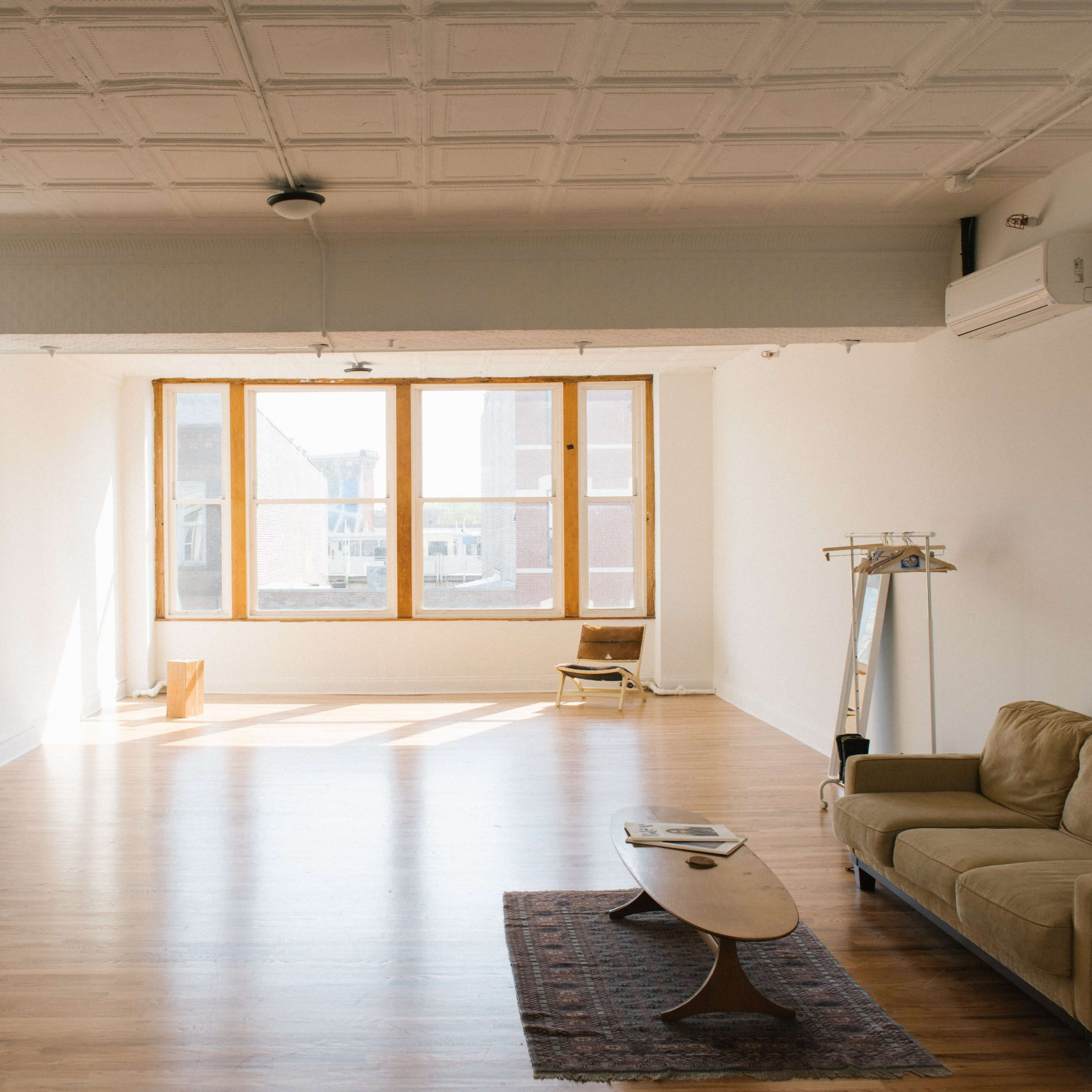 A spacious, brightly lit room with a wooden floor, large windows, a beige sofa, a low coffee table, and a dining area with black chairs.