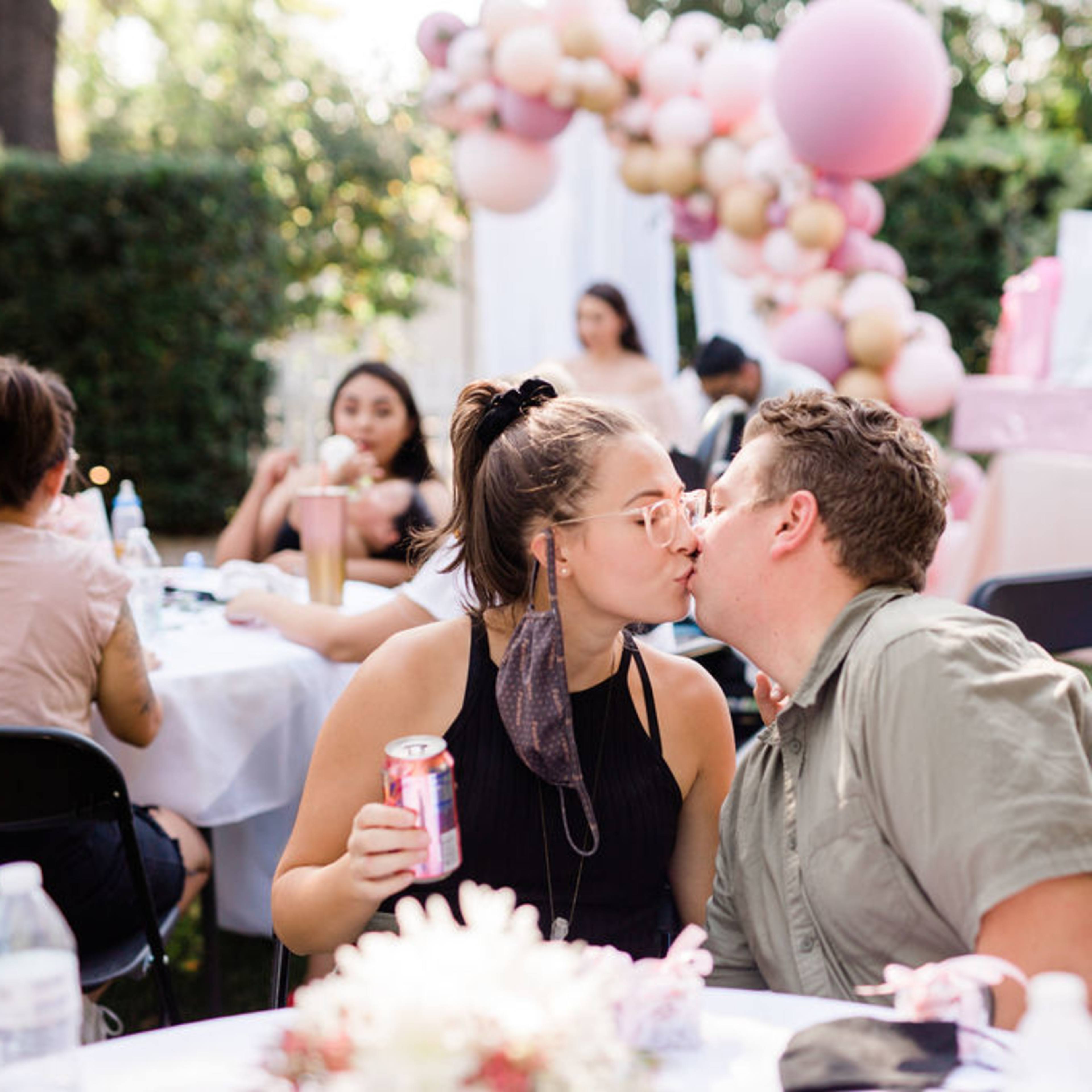 A couple shares a kiss at a celebration with balloons and gifts in the background while seated at a table.