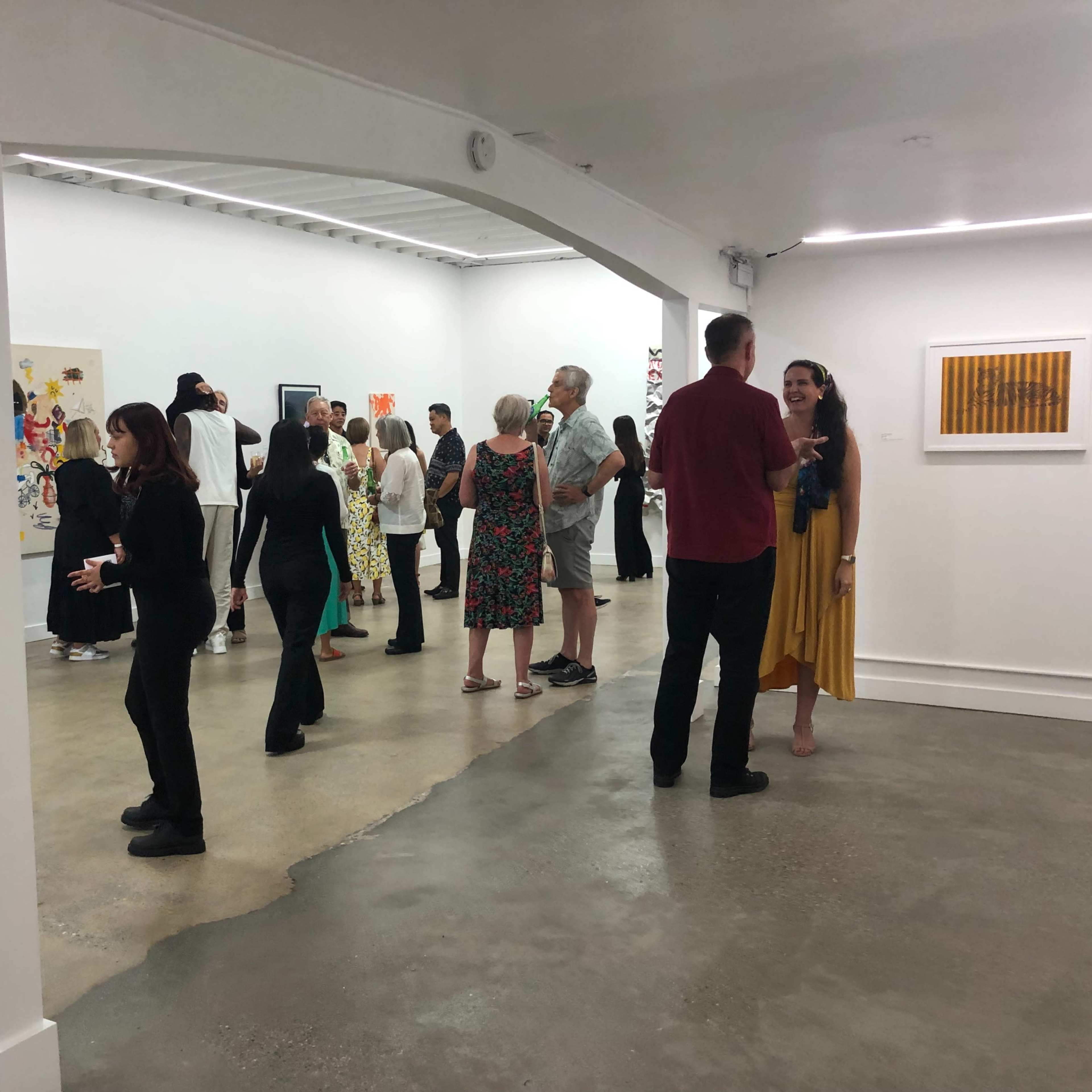 A group of people is gathered in a spacious art gallery, engaging in conversation while observing various artworks on the walls.