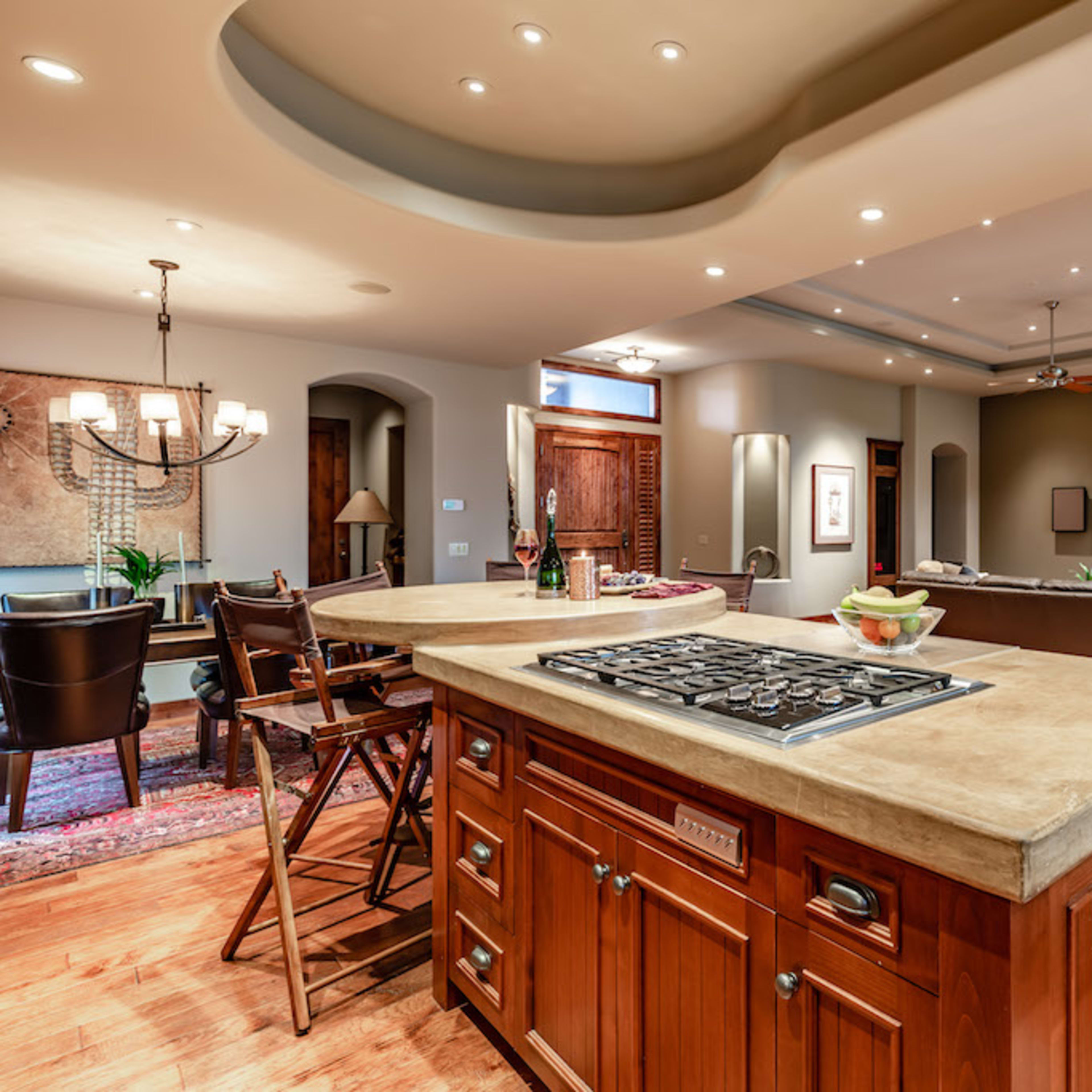 A spacious kitchen features a central island with a stovetop, surrounded by dining and living areas with wooden flooring and recessed lighting.