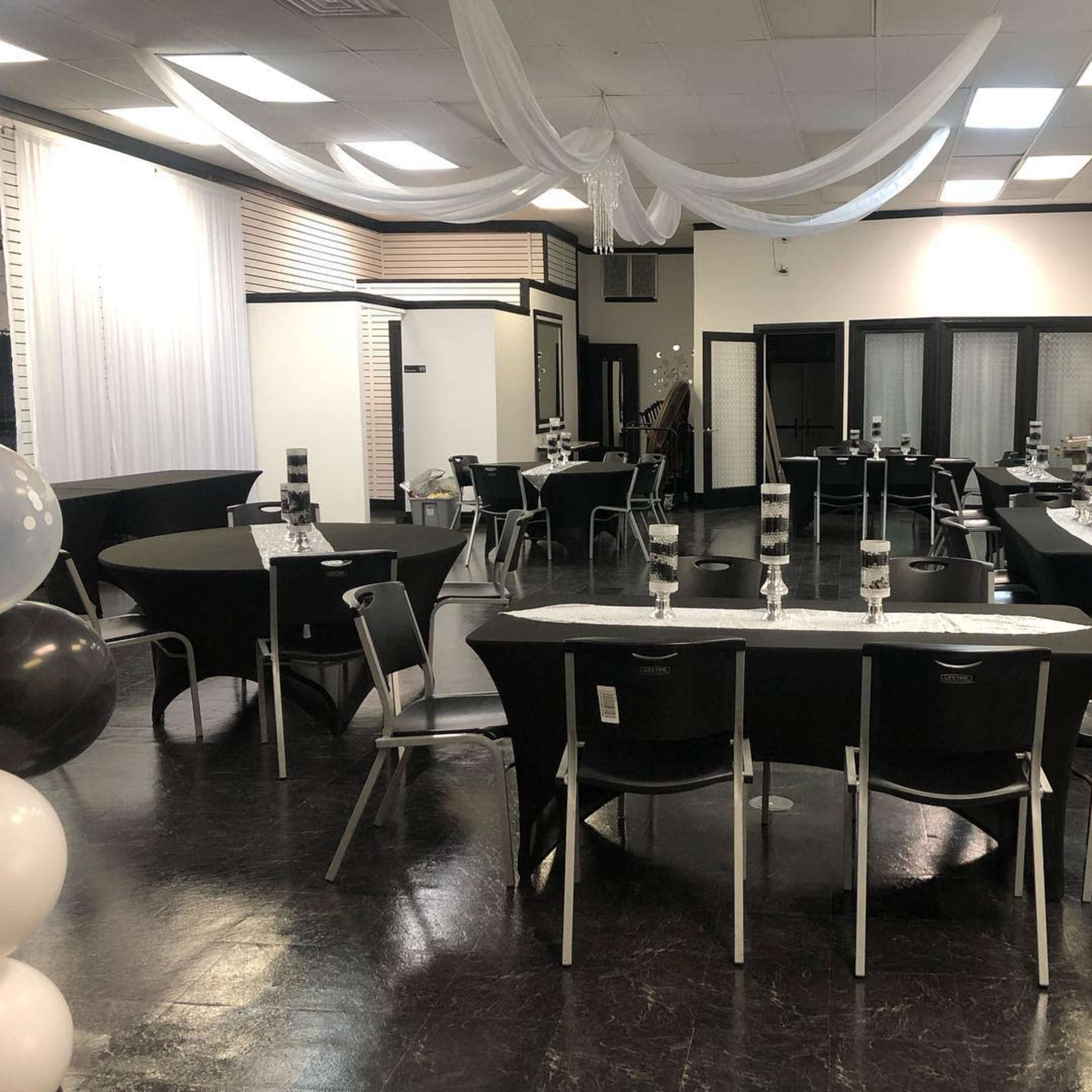 A large event space is set up with several round and rectangular tables, each adorned with tall glass centerpieces, and draped fabric hangs from the ceiling.