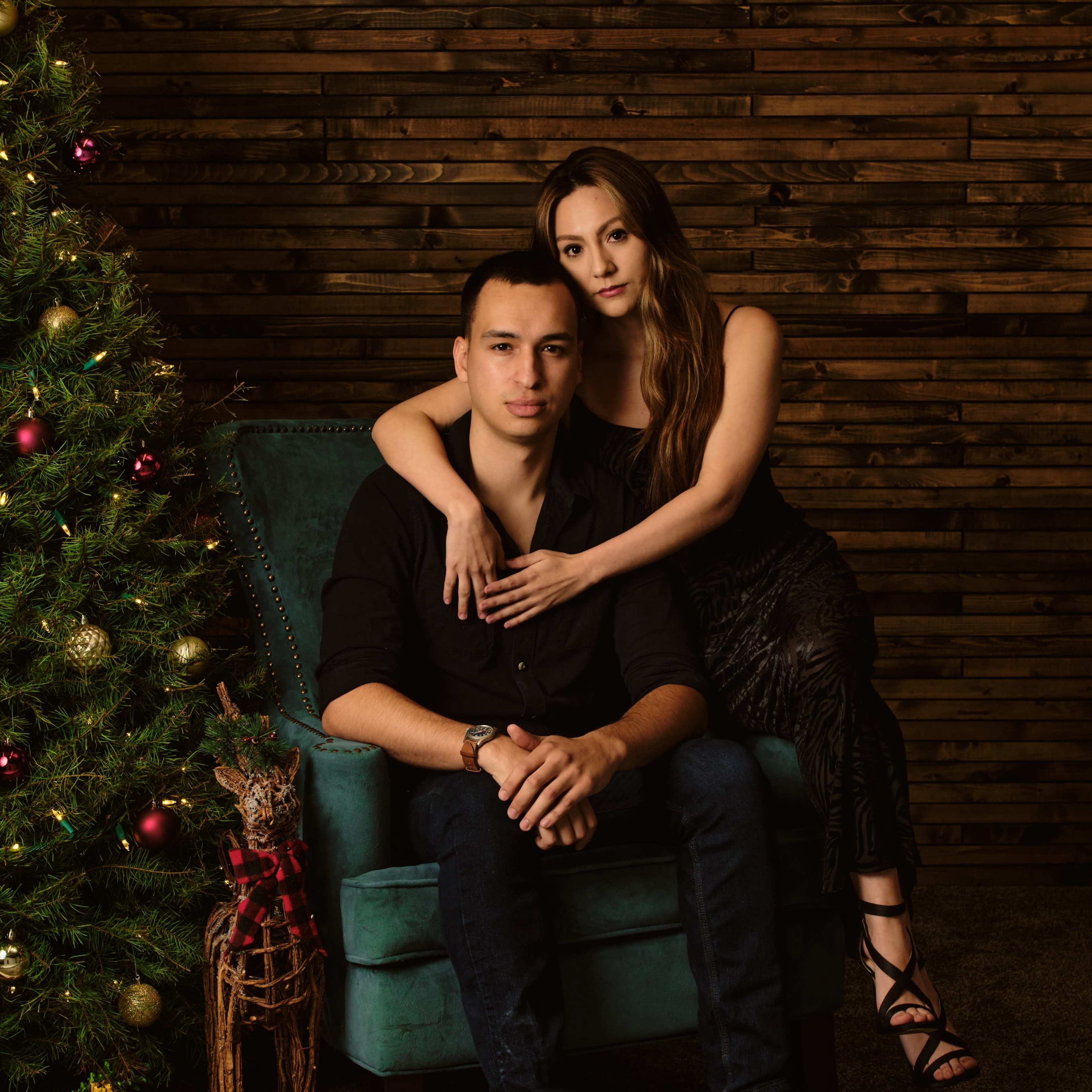 A couple poses together on a teal chair beside a decorated Christmas tree in a room with wooden walls.