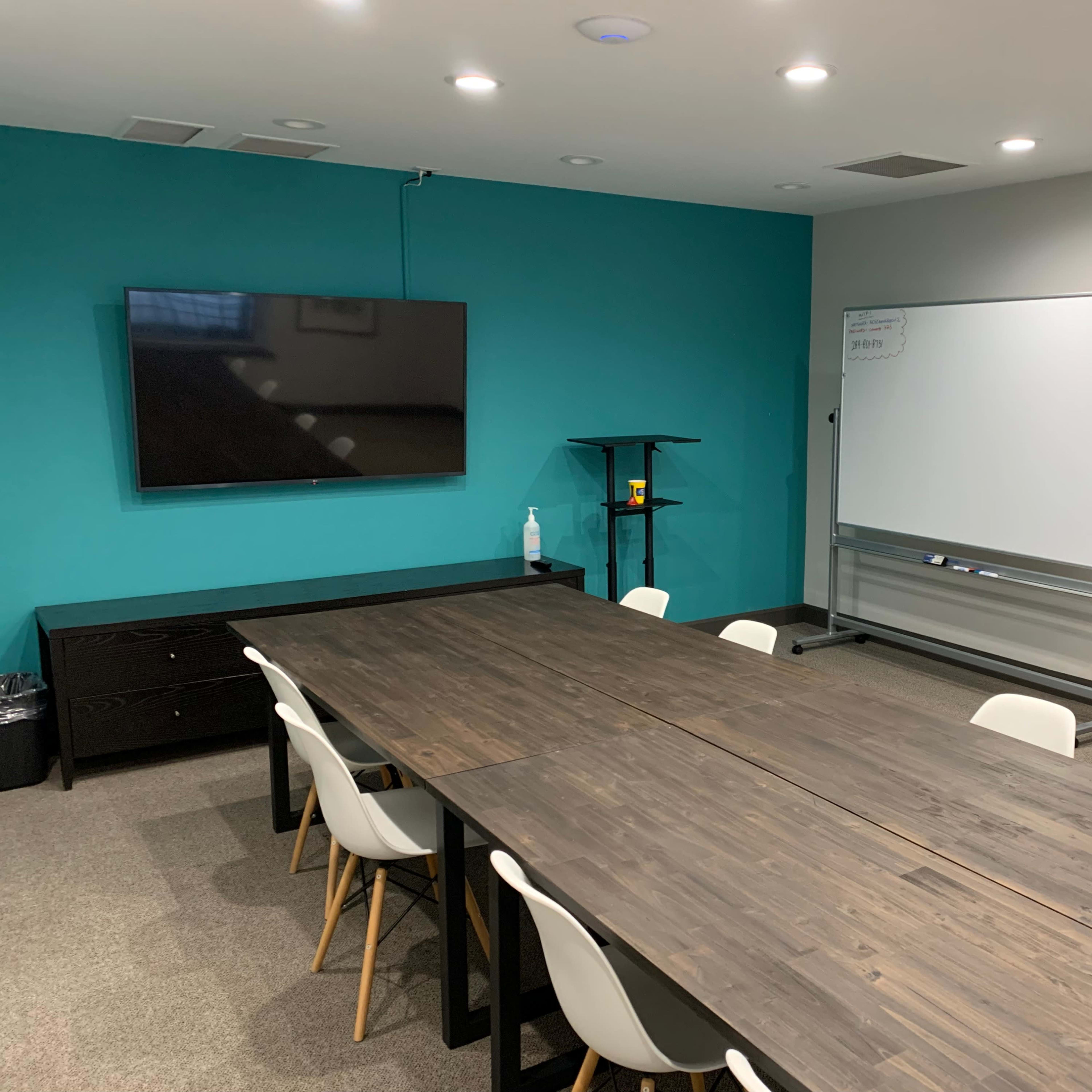 A conference room with a large wooden table, white chairs, a wall-mounted TV, and a whiteboard on a blue wall.