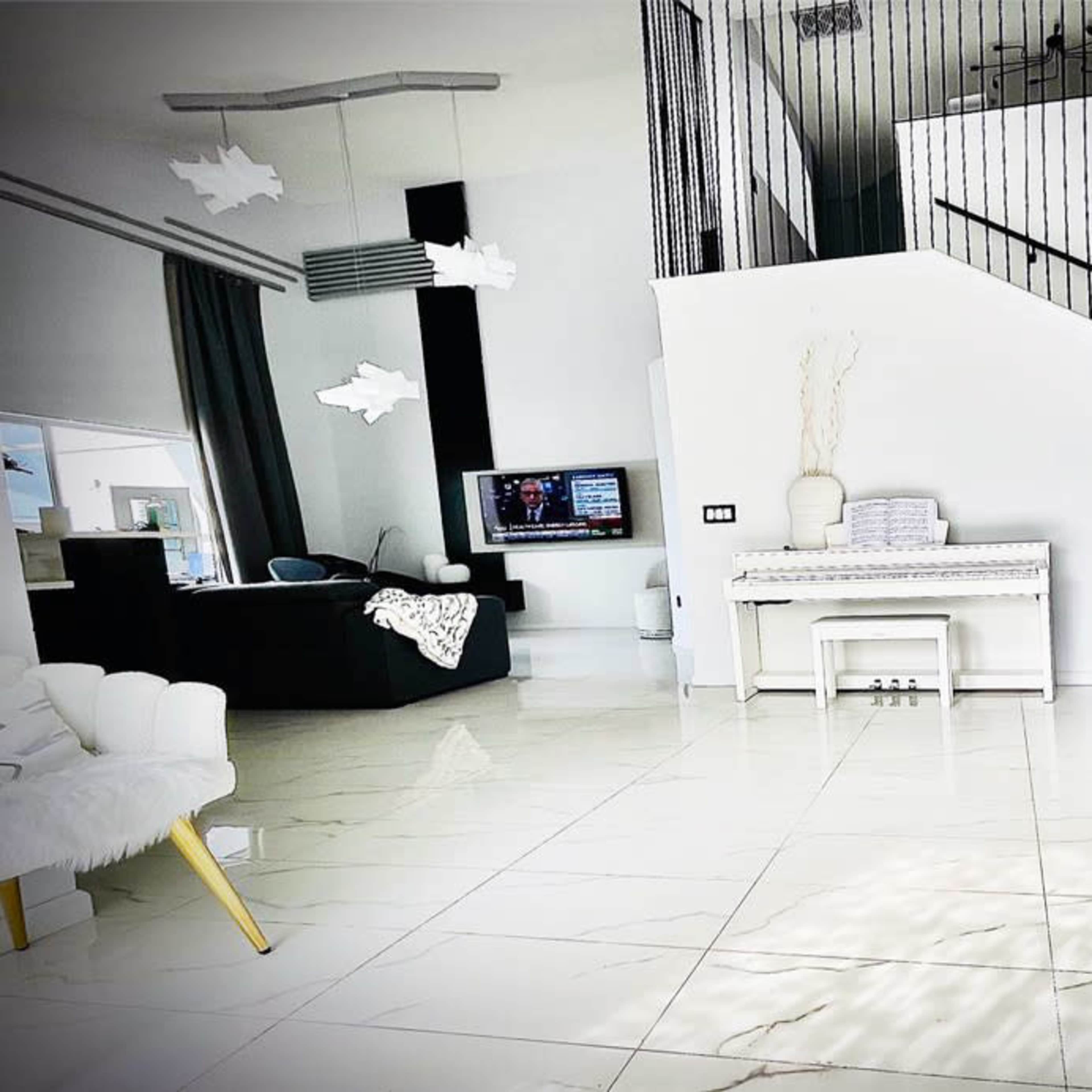 A modern, minimalist interior featuring a staircase, a white piano, and contemporary furniture, with large windows allowing natural light.