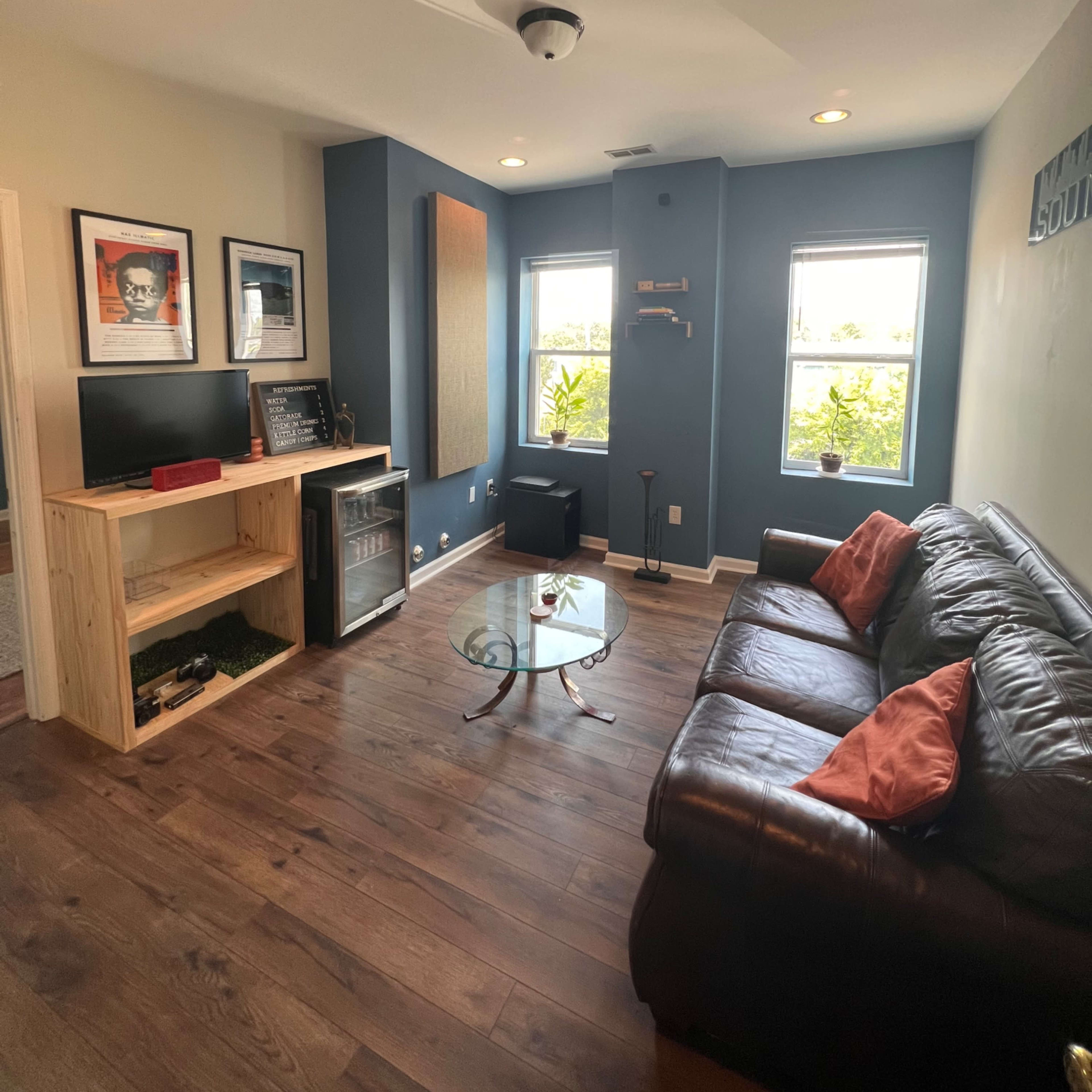 A small living room with a black leather sofa, a glass coffee table, a wooden shelf unit, a television, and large windows allowing natural light.