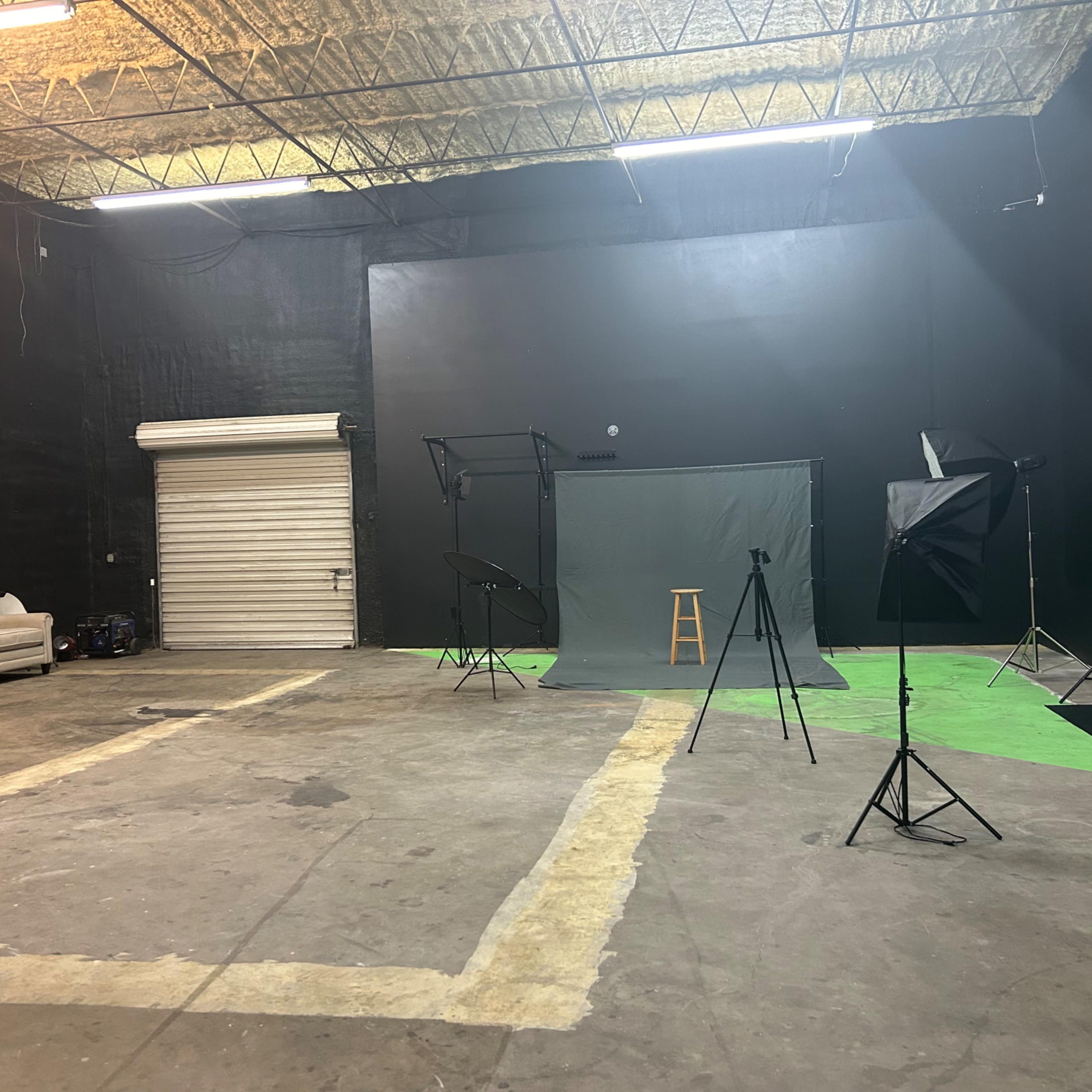 A spacious, industrial studio with black walls, various lighting equipment, and a green backdrop set up for photography or filming.