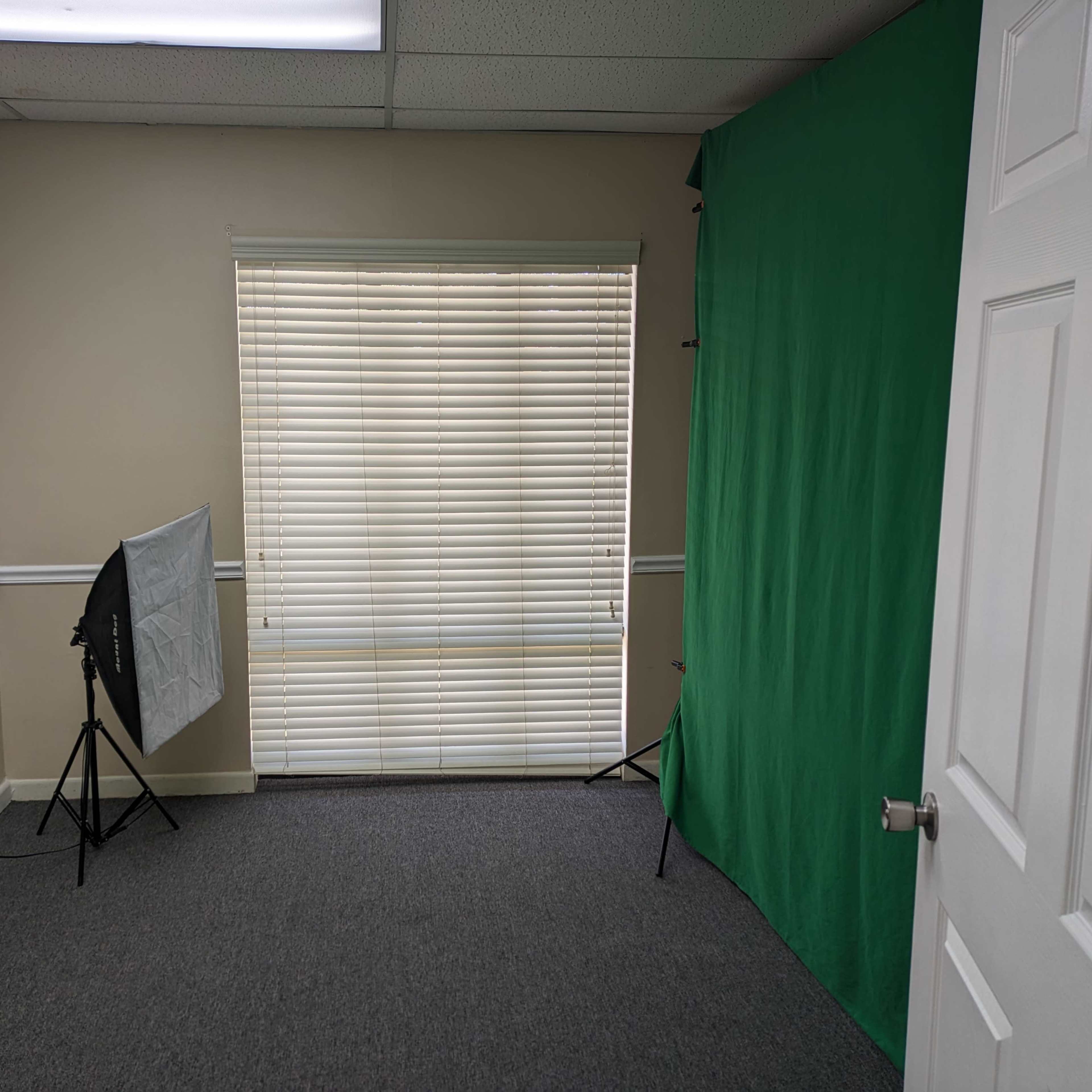 A small room with a green backdrop, a window with blinds, and two softbox lights set up on stands.