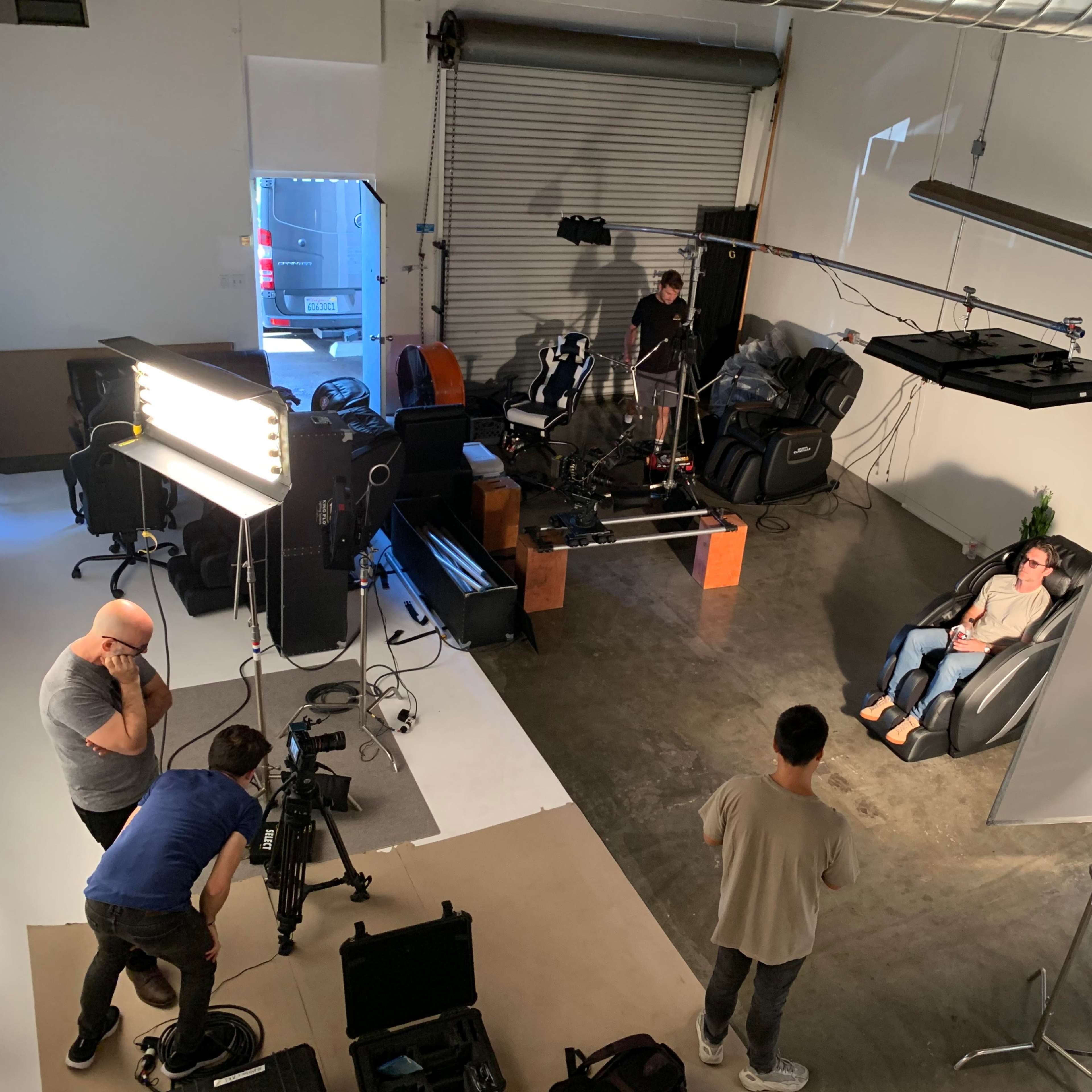 A film crew sets up equipment in a studio, featuring a chair and multiple cameras while a person sits in the chair.