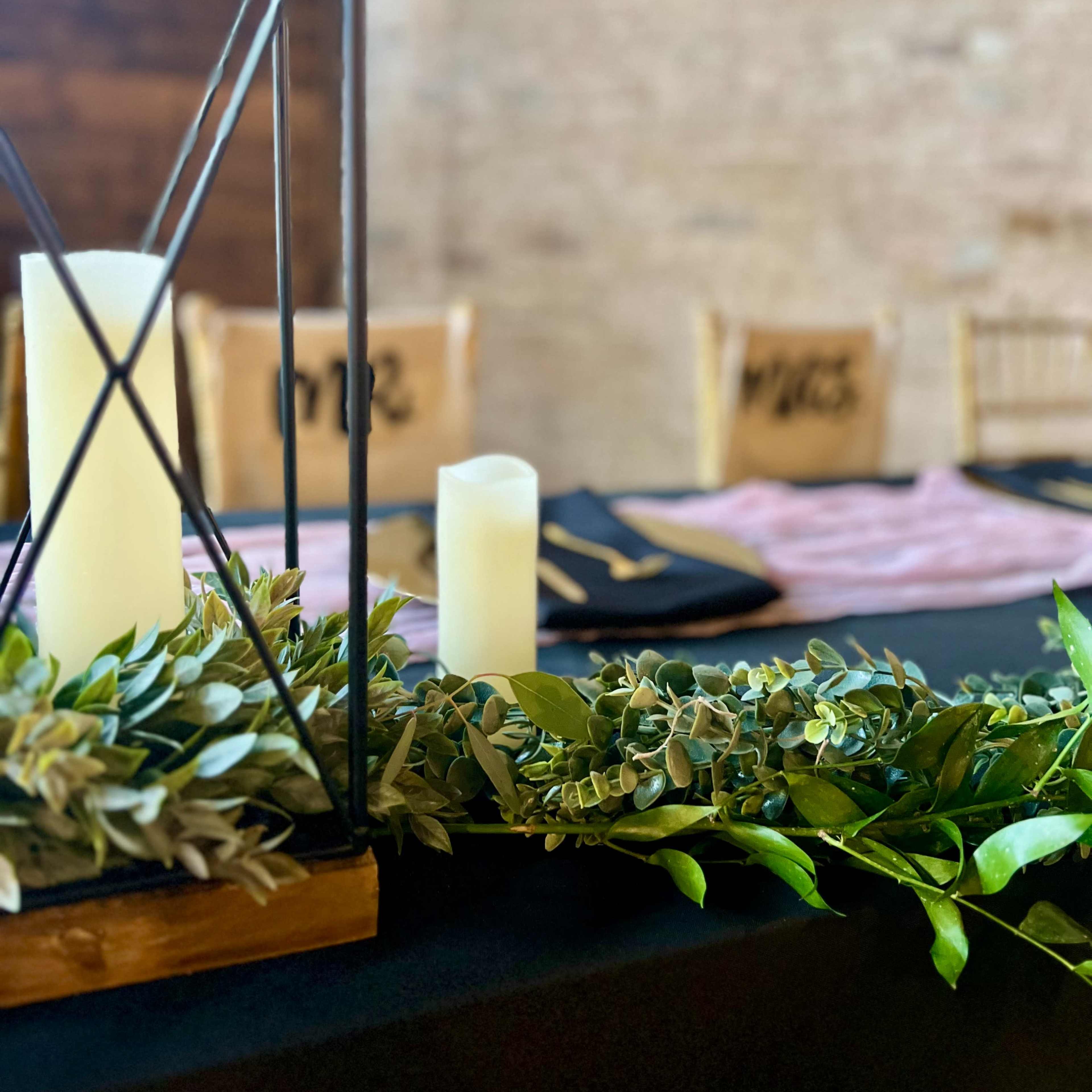 A table setting features a geometric lantern, candles, and a garland of greenery against a backdrop of burlap-covered chairs.