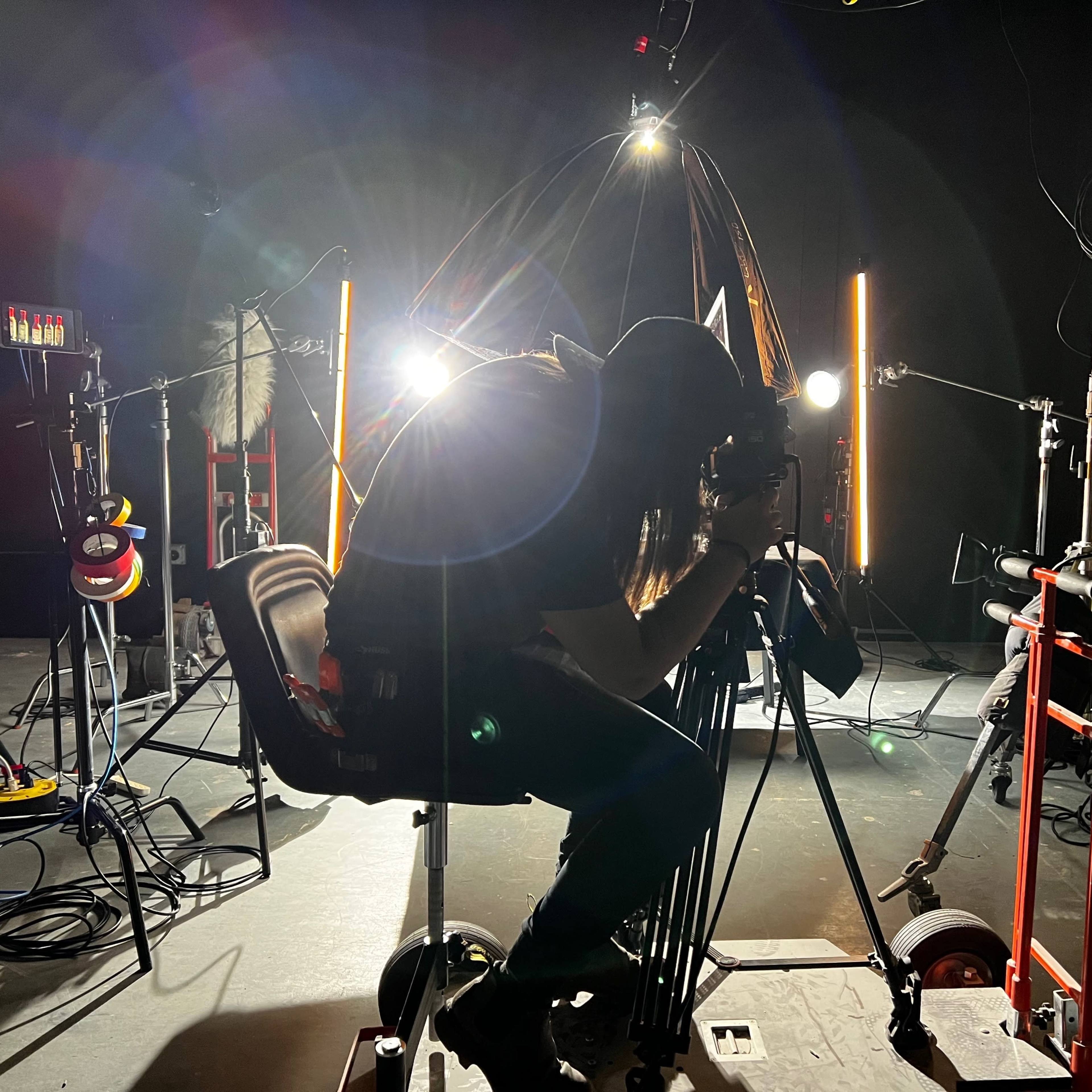 A photographer crouches on a chair while adjusting a camera amidst studio equipment and bright lighting.