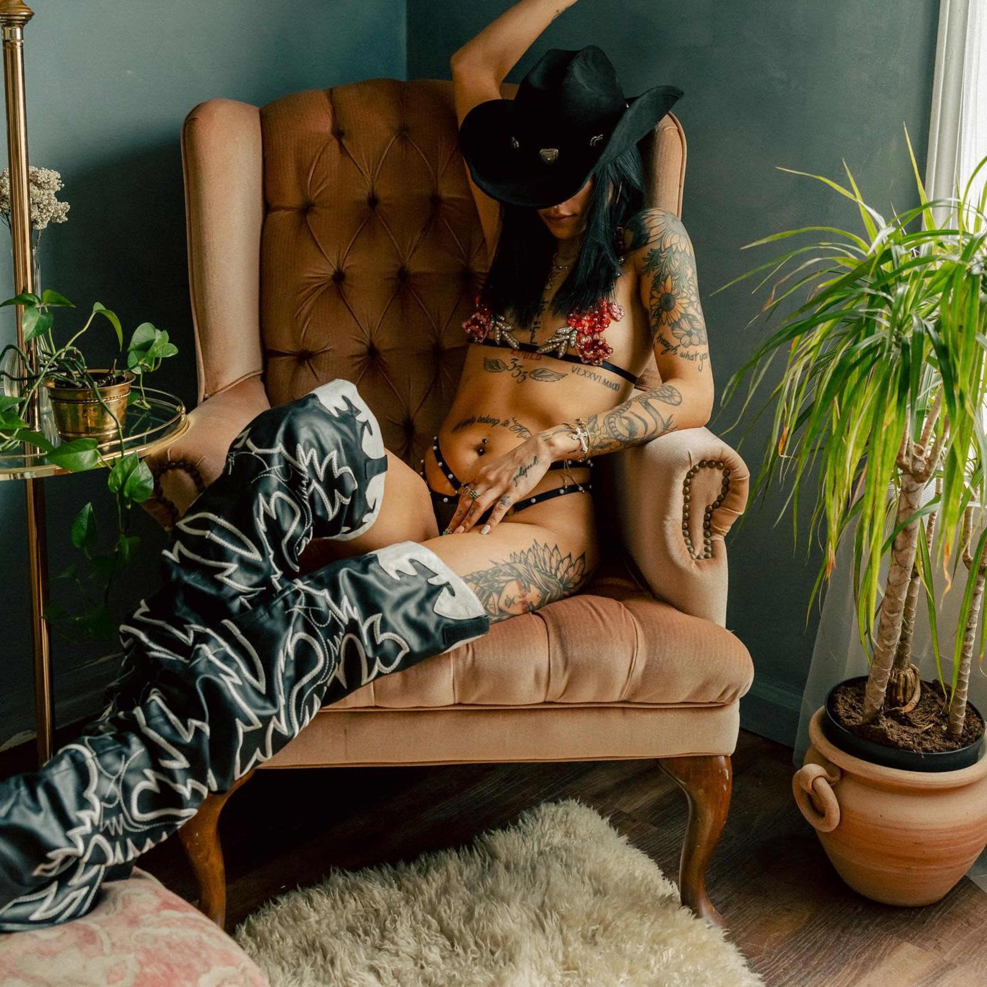 A person with tattoos sits in a vintage armchair, wearing lingerie and tall patterned boots, in a room with indoor plants and decorative wall art.