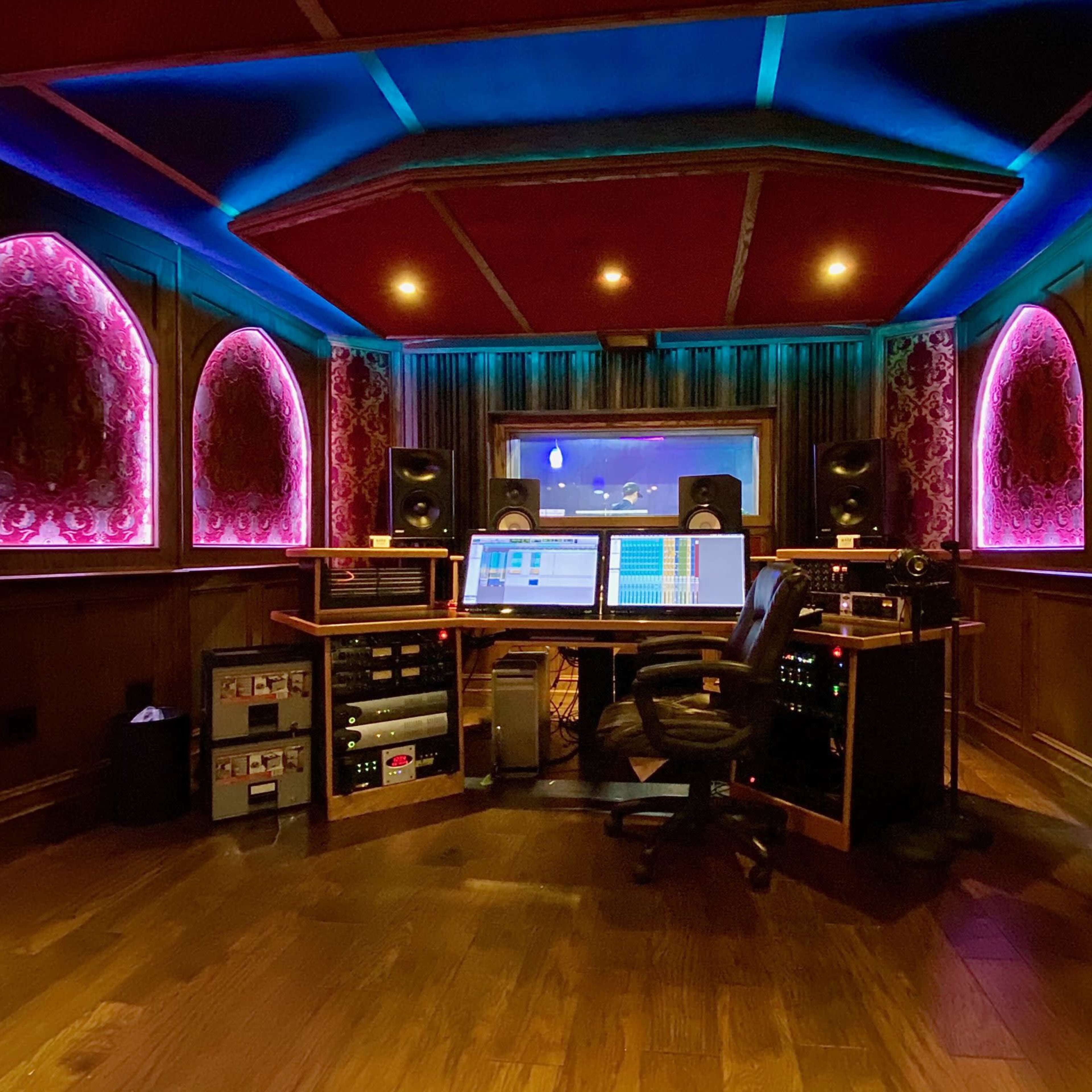 A modern recording studio with colorful lighting, a central mixer, and sound equipment arranged along wooden walls.
