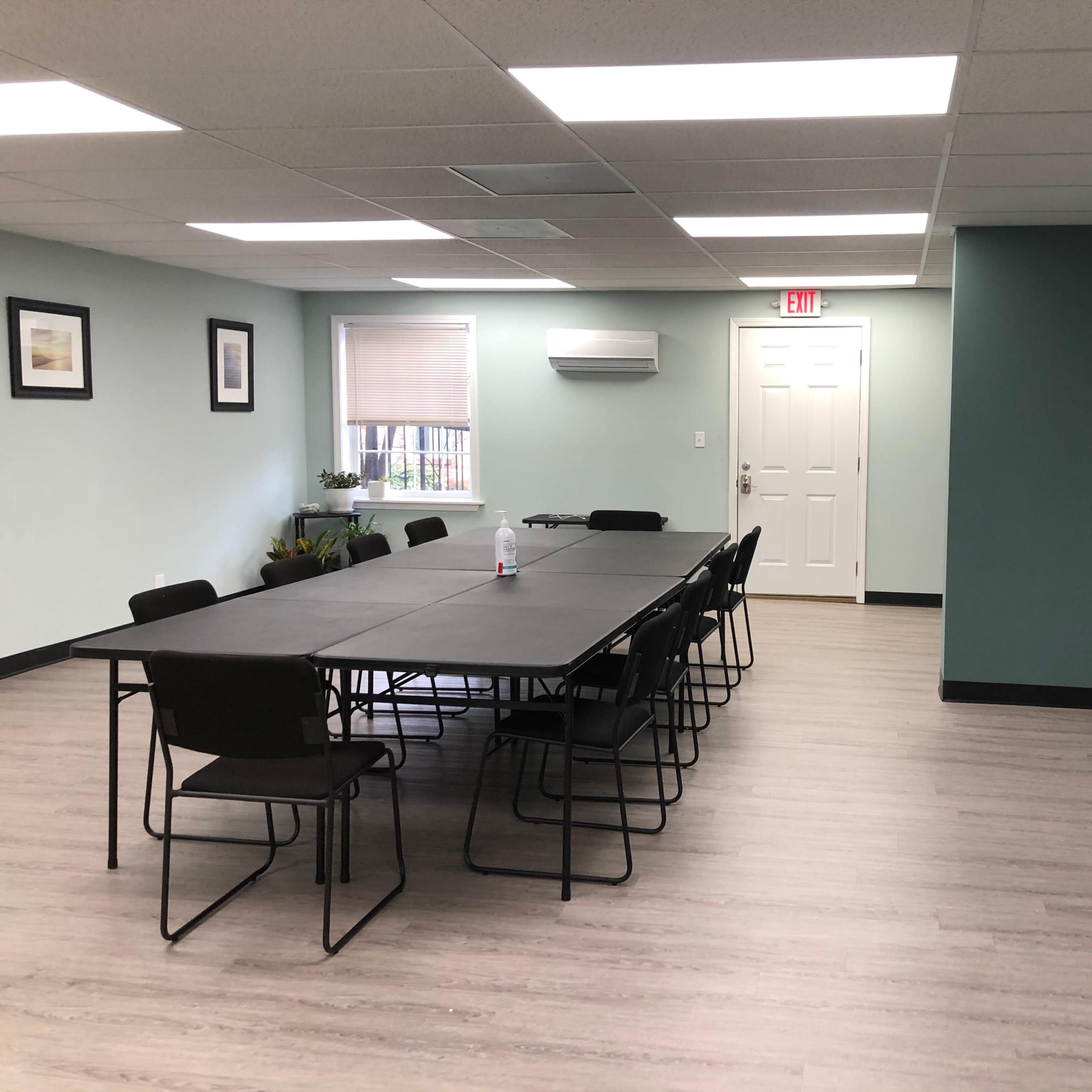 A spacious meeting room with a long table surrounded by black chairs, a door at the back, and a television mounted on the wall.