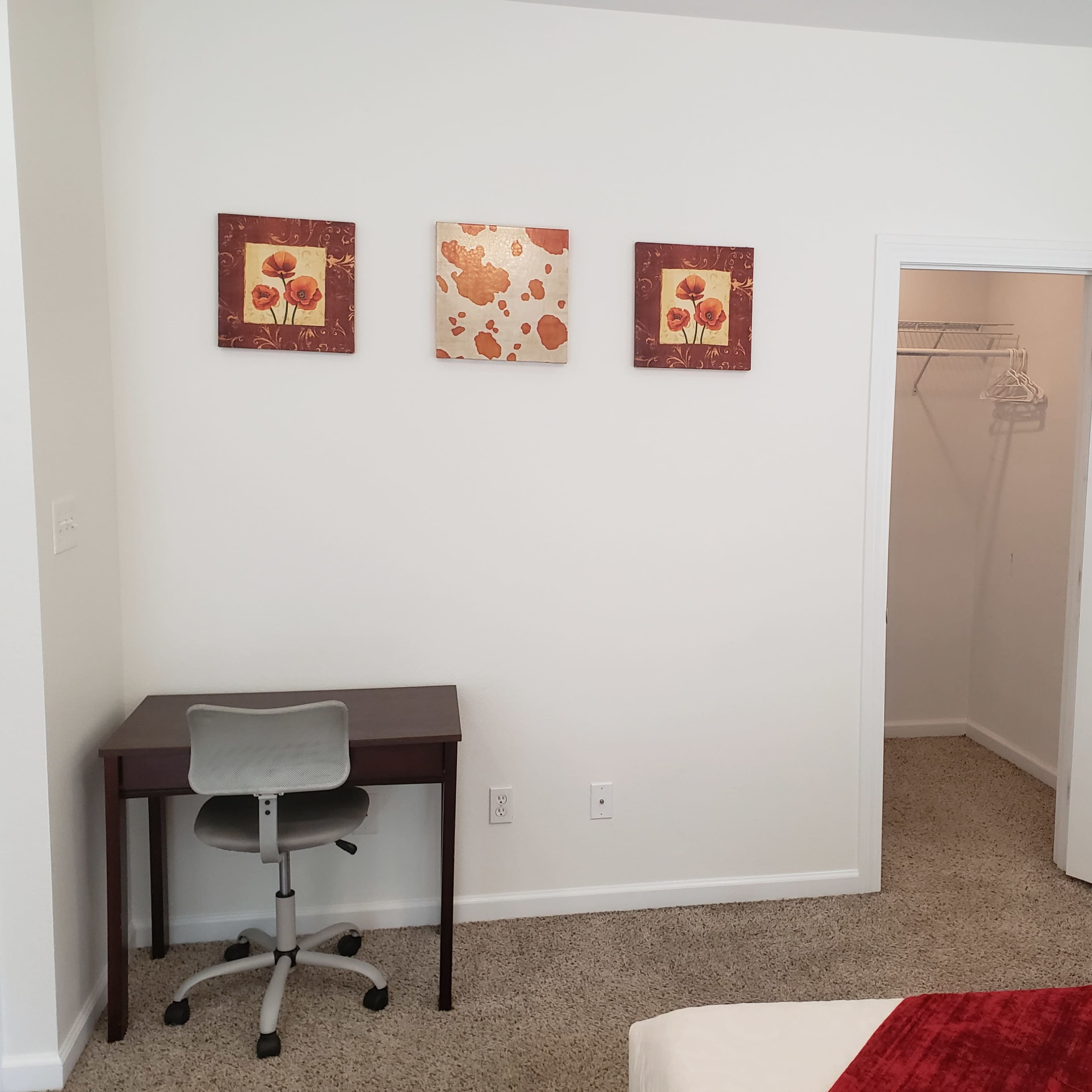 A room with a desk and three floral wall art pieces, next to an open closet containing hangers.