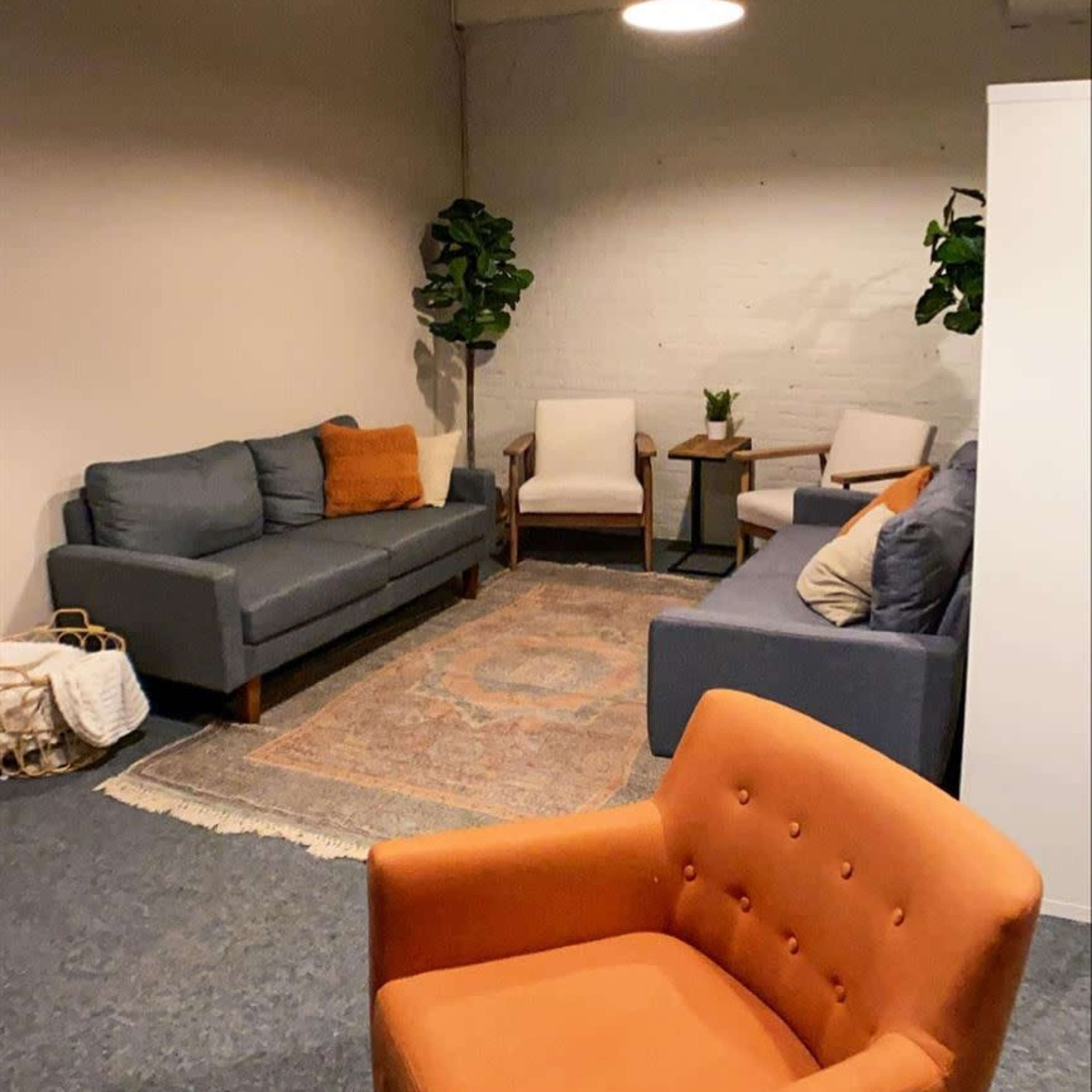 A cozy lounge area with gray sofas, an orange armchair, a small side table, and green plants in a softly lit room.