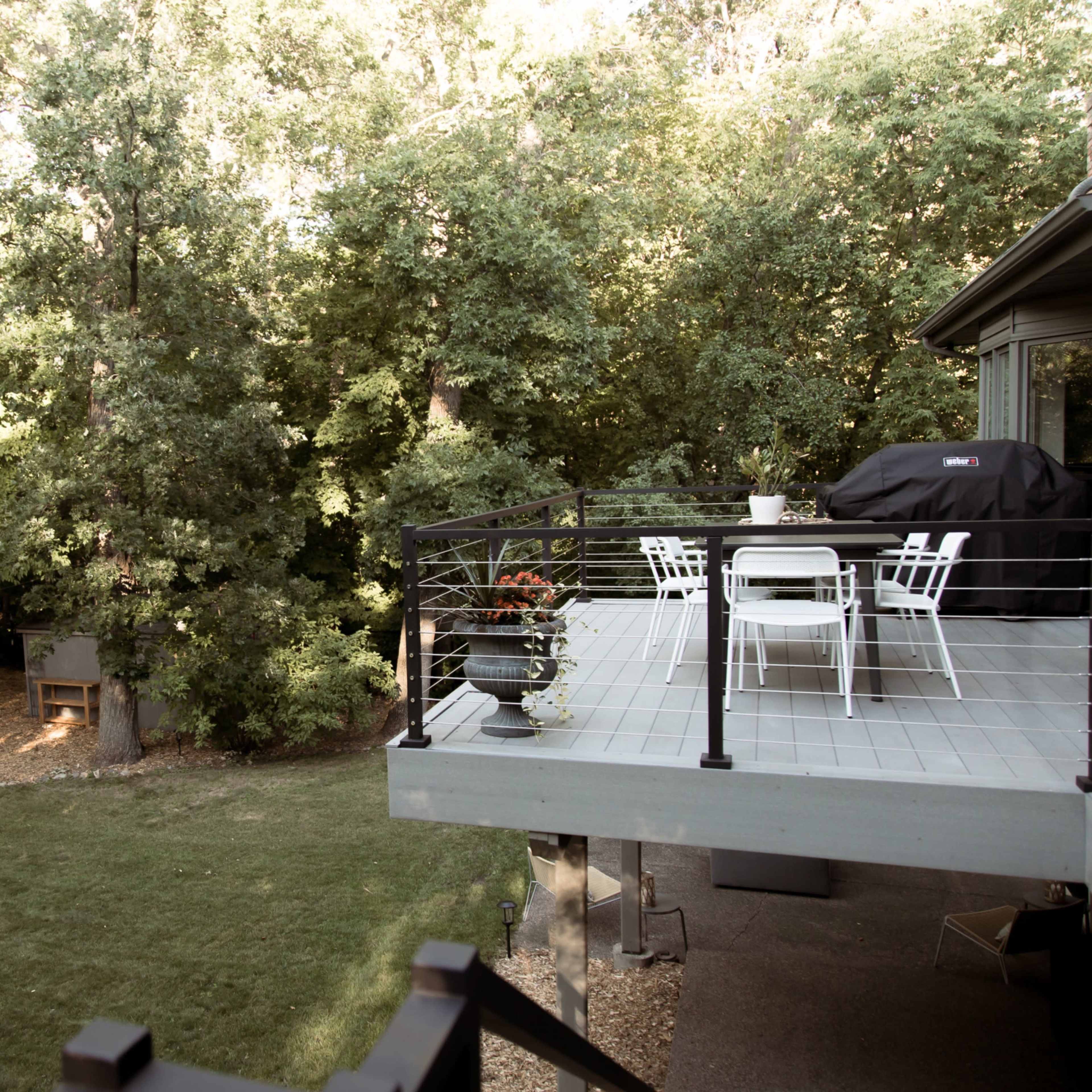 A wooden deck with white patio furniture overlooks a grassy area surrounded by trees.