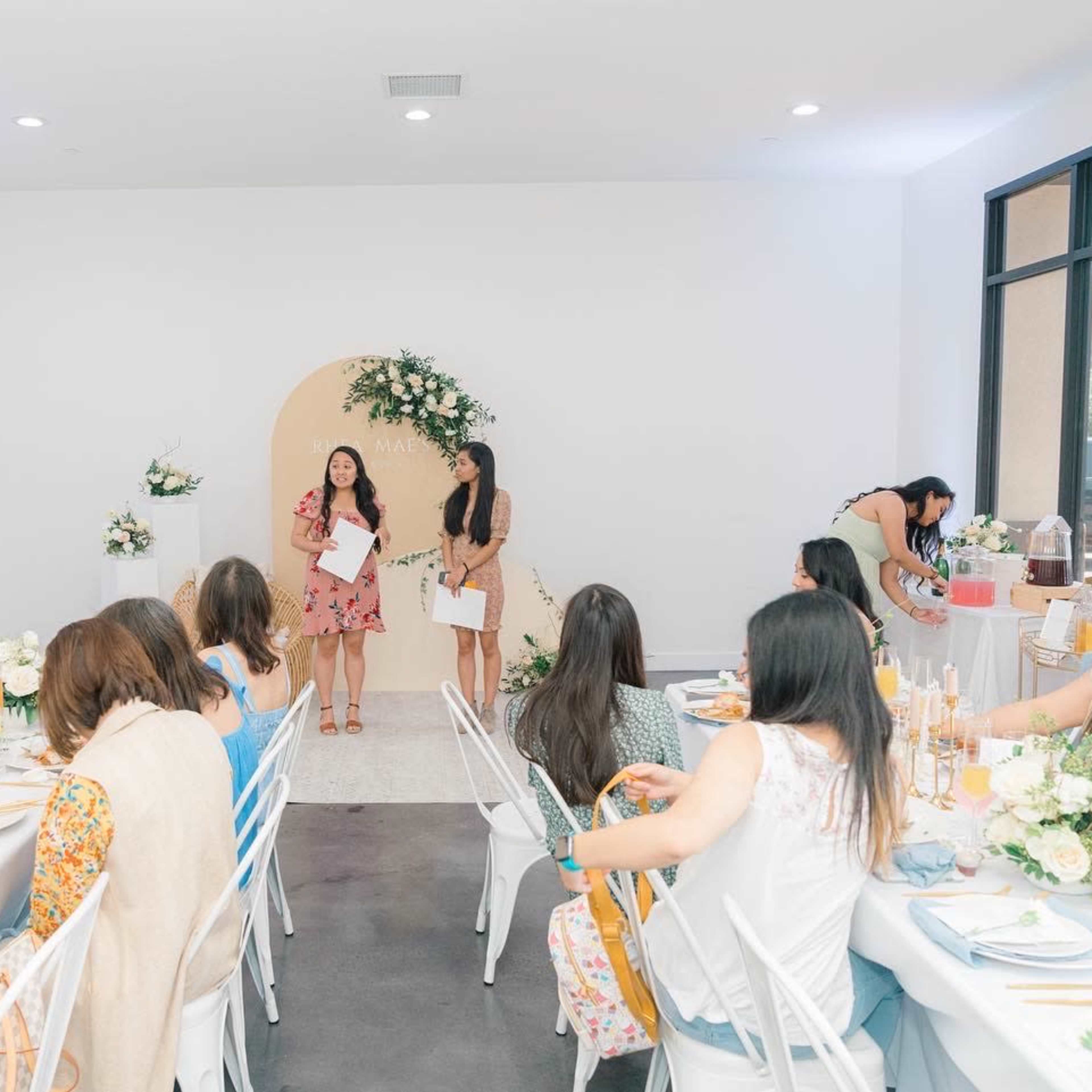 A group of women sit at elegantly set tables while two speakers present in front of a floral backdrop in a bright, modern event space.