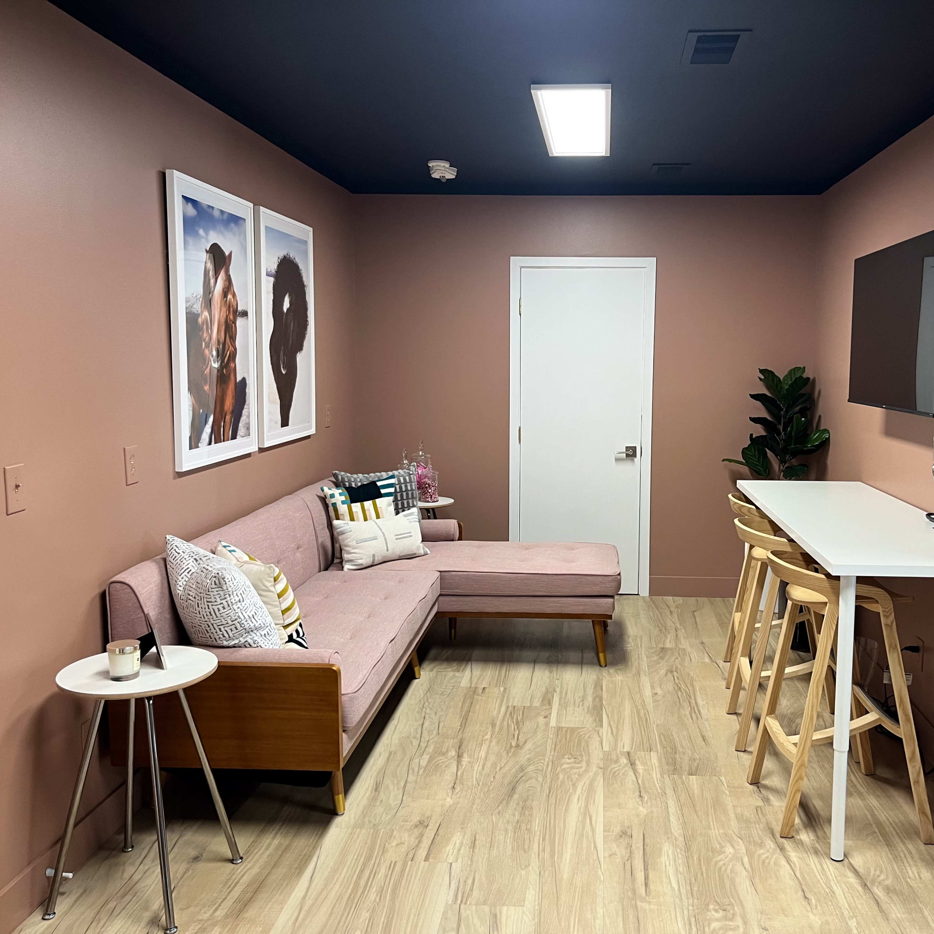 A modern living space with a pink wall, a pink couch, a white table with bar stools, and a large mirror next to a television.