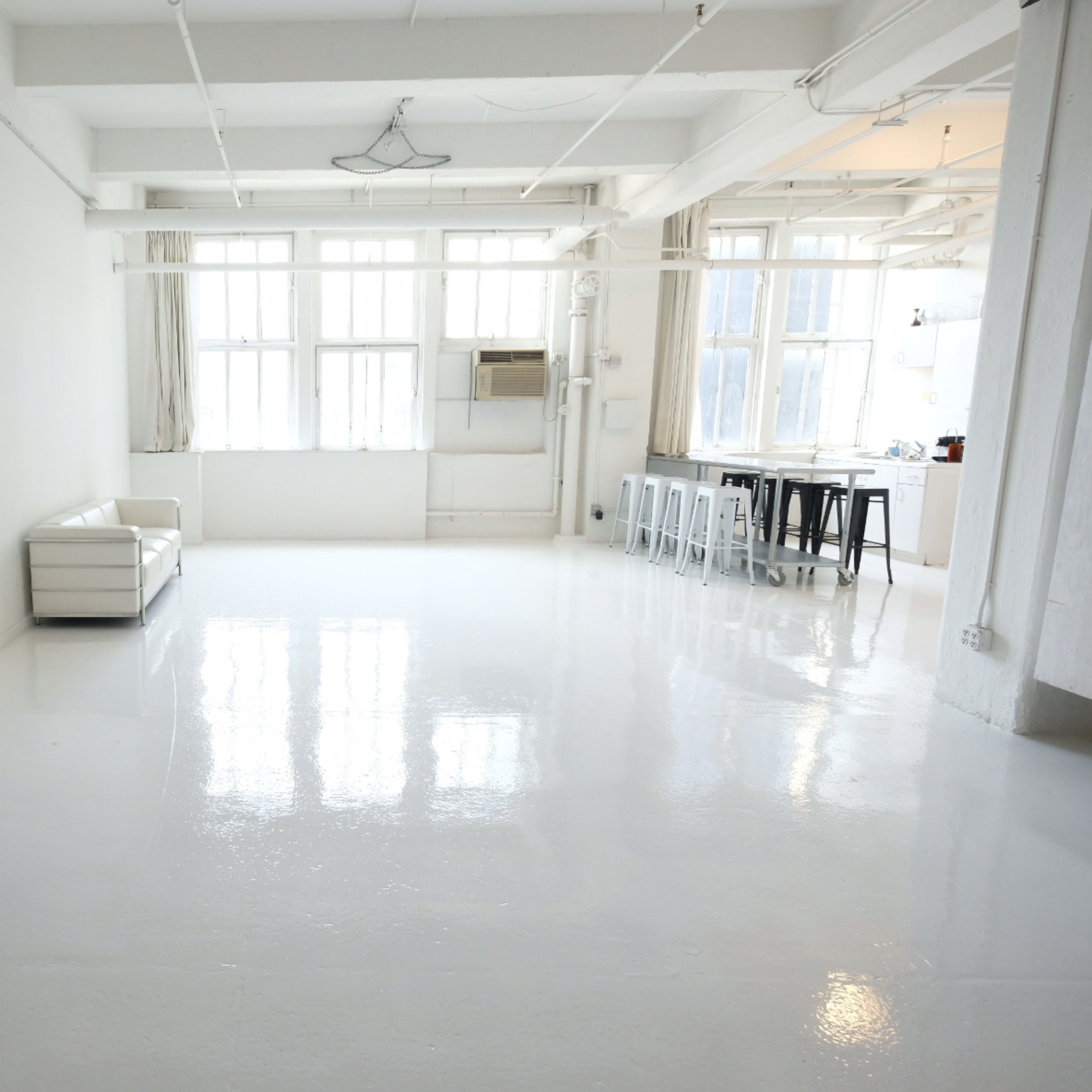 A spacious, minimalist interior with white walls and glossy flooring, featuring a white sofa, a metallic table on wheels, and a kitchen area in the background.