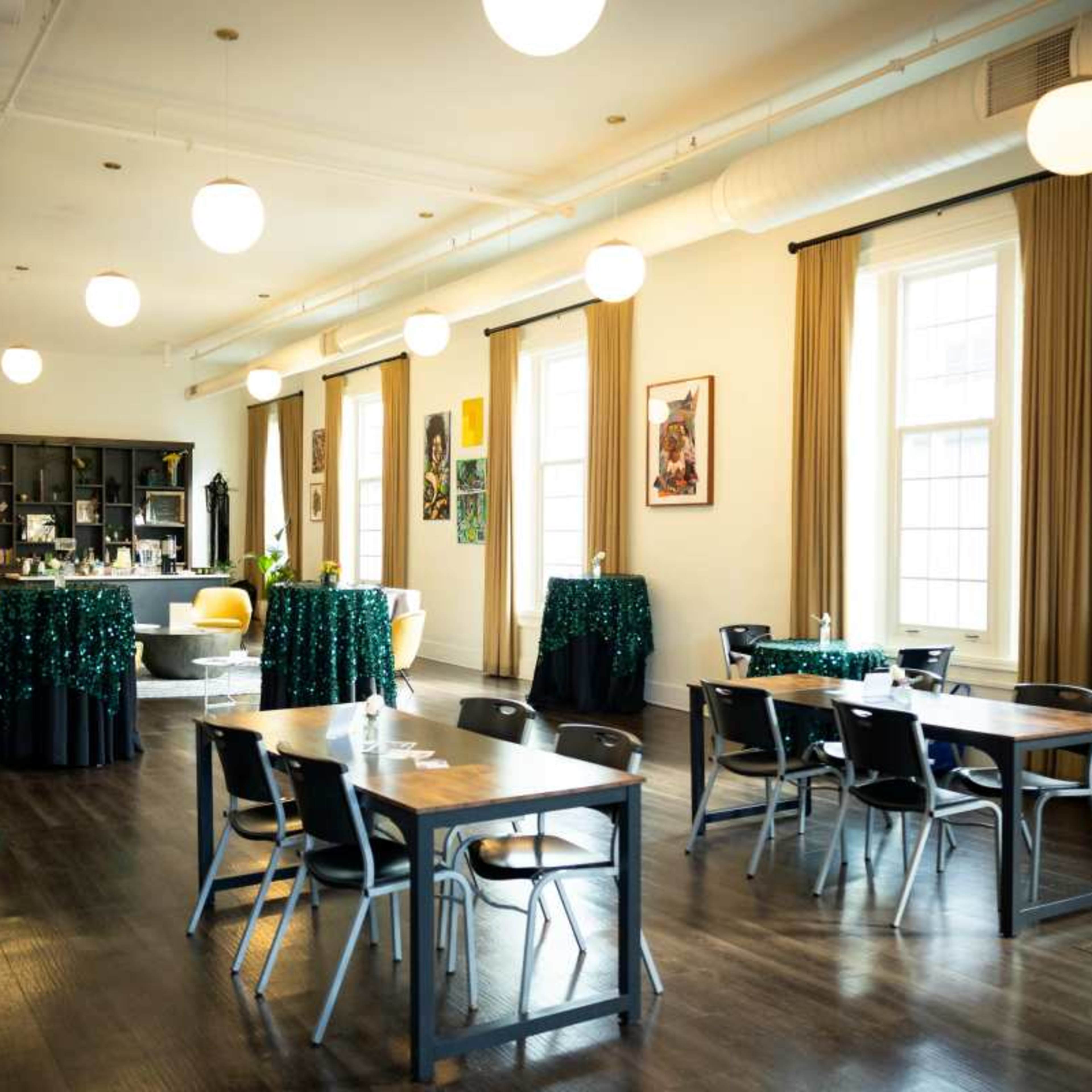 A bright, spacious event space with tables and chairs arranged, green sequin tablecloths, and large windows along the walls.