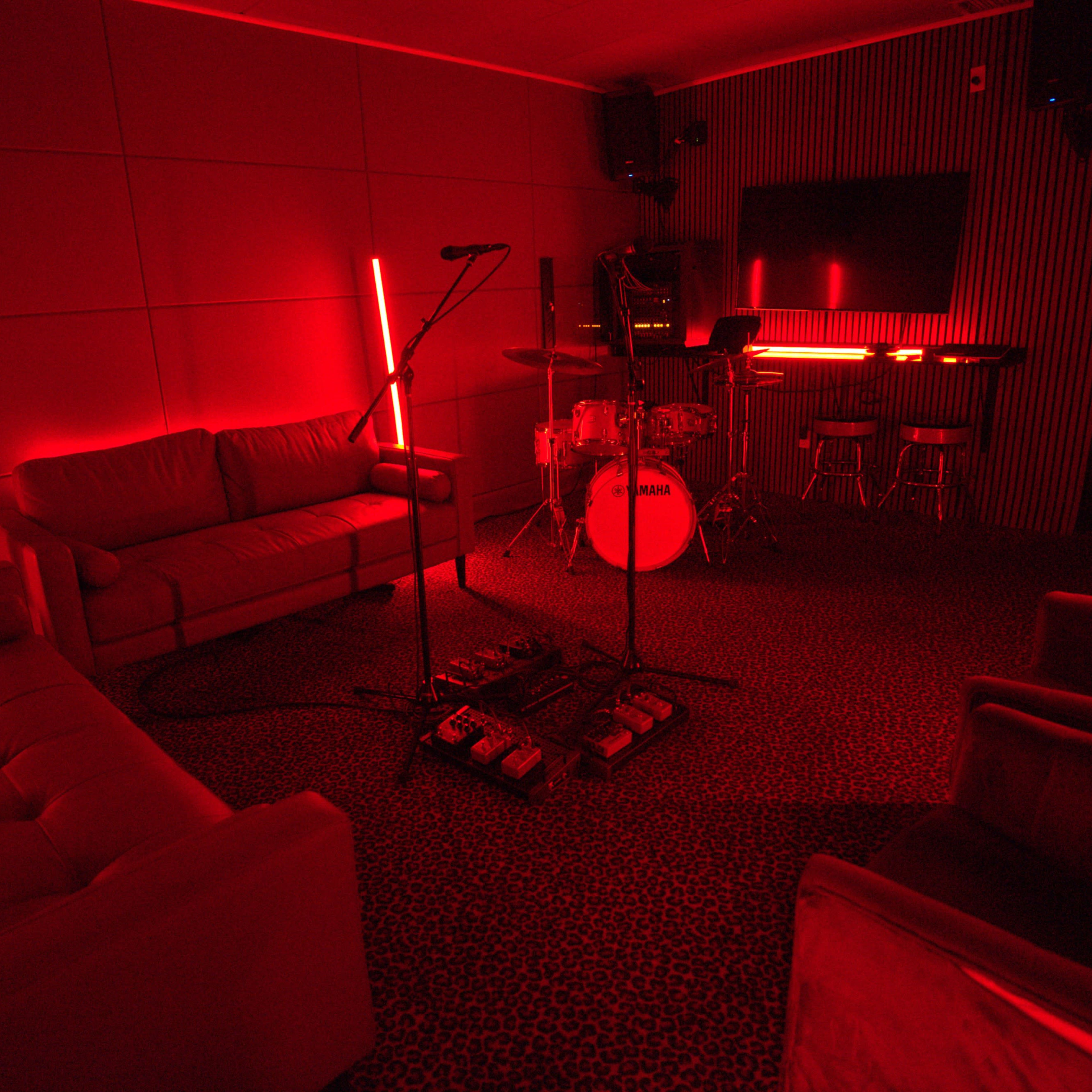 A music practice room featuring red lighting, a microphone, drums, and comfortable seating.