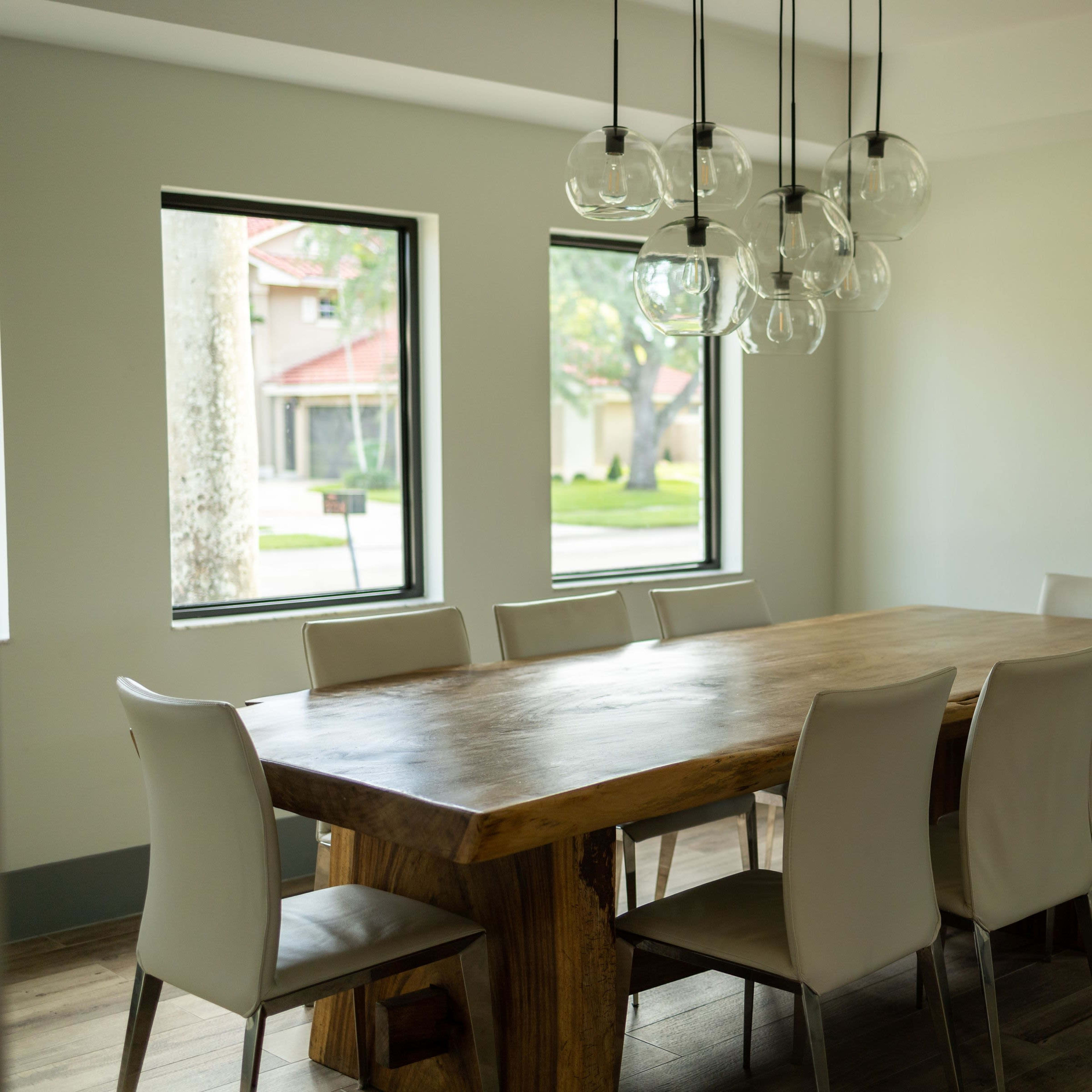A wooden dining table with eight light-colored chairs is set in a brightly lit room featuring large windows and modern pendant lighting.
