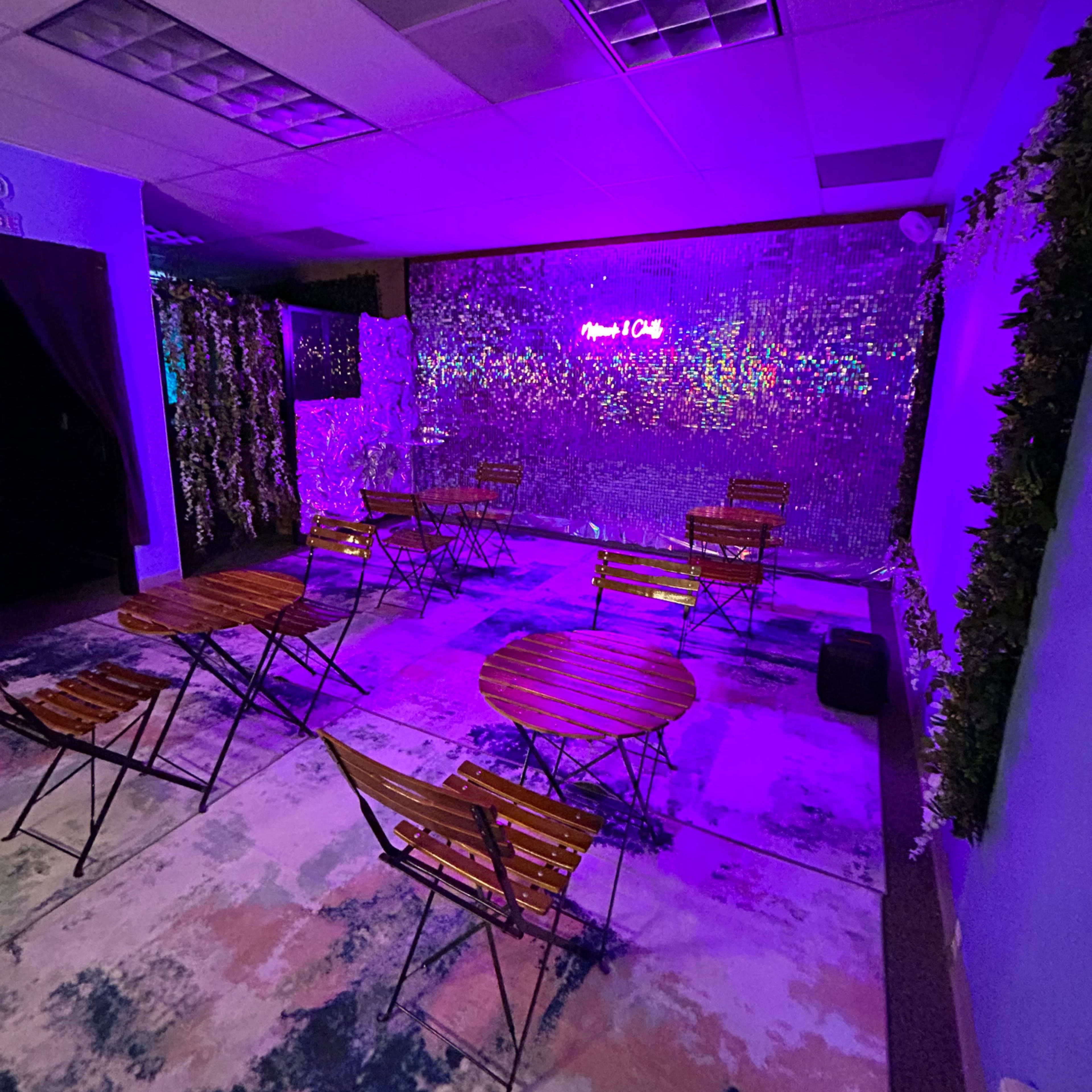 A cozy event space features several metal bistro tables and chairs arranged on a colorful rug, with a shimmering backdrop illuminated by purple lights.
