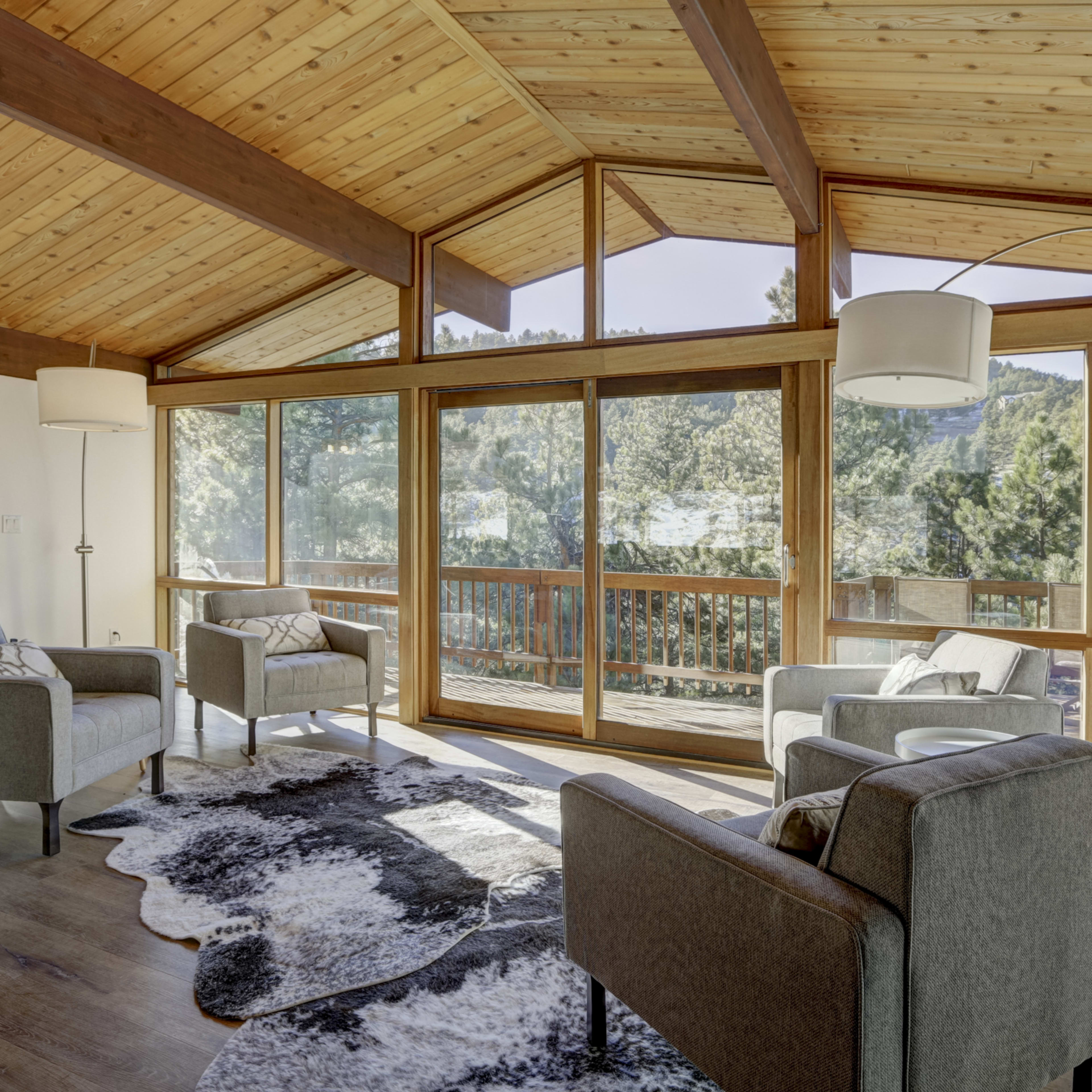 A spacious living room features large windows overlooking a deck and trees, with modern seating arranged on a patterned rug.