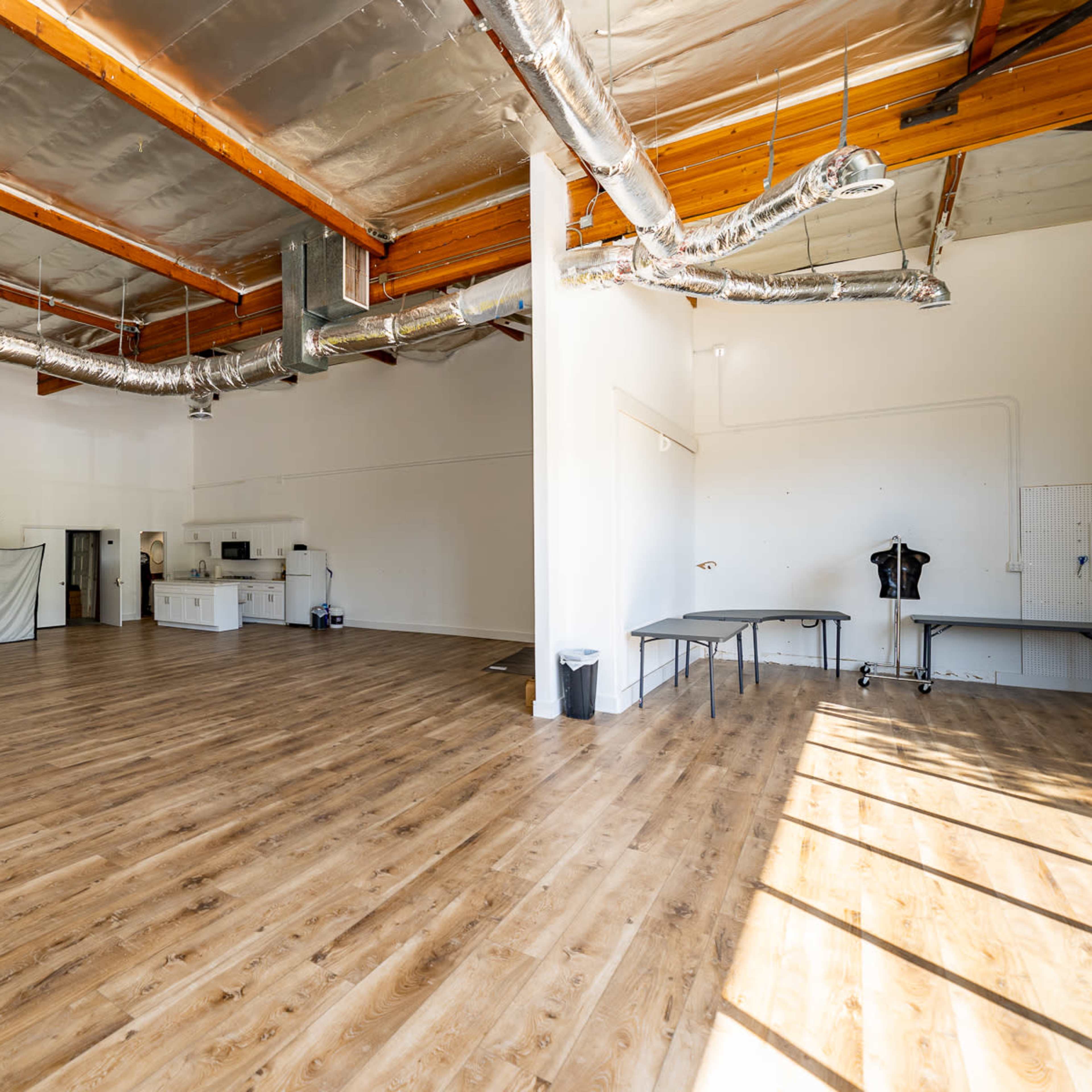 A spacious photography studio with wooden floors, white walls, and various equipment set up, including a stool and tables.