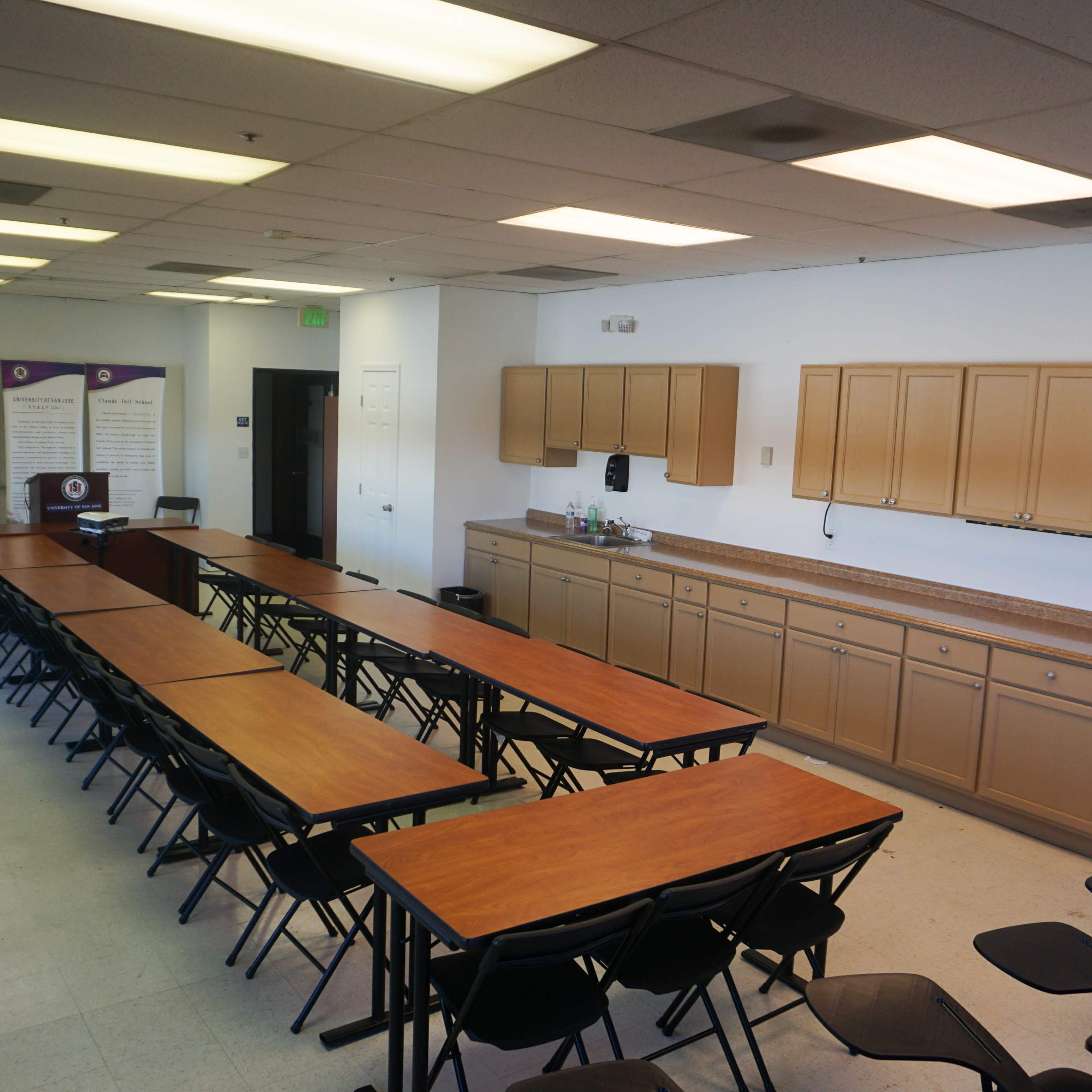 A meeting room features several rows of folding tables and chairs, with a kitchenette along one wall and a projector screen set up in the corner.