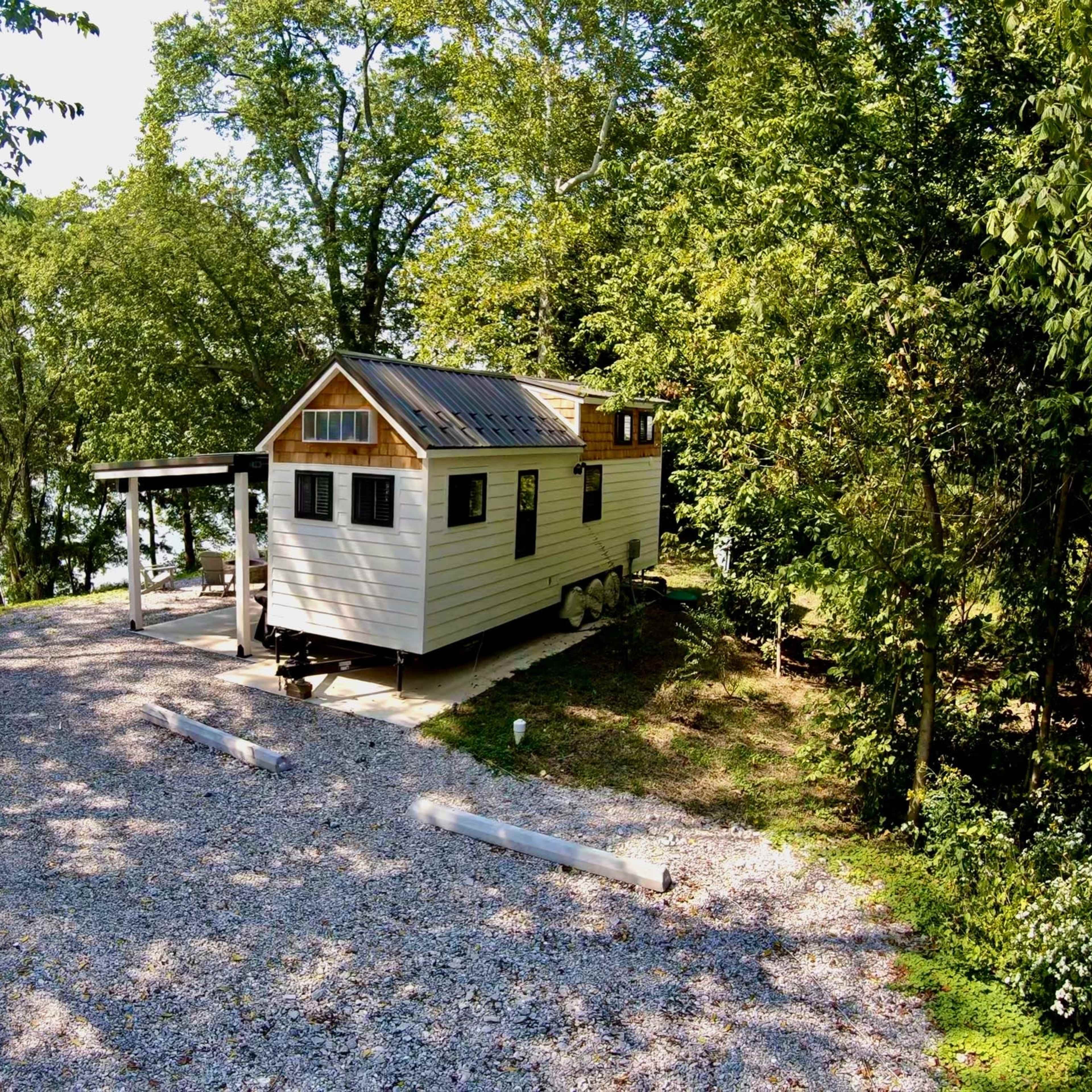 A small cabin with a sloped roof is parked on a gravel driveway beside a lake, surrounded by trees.