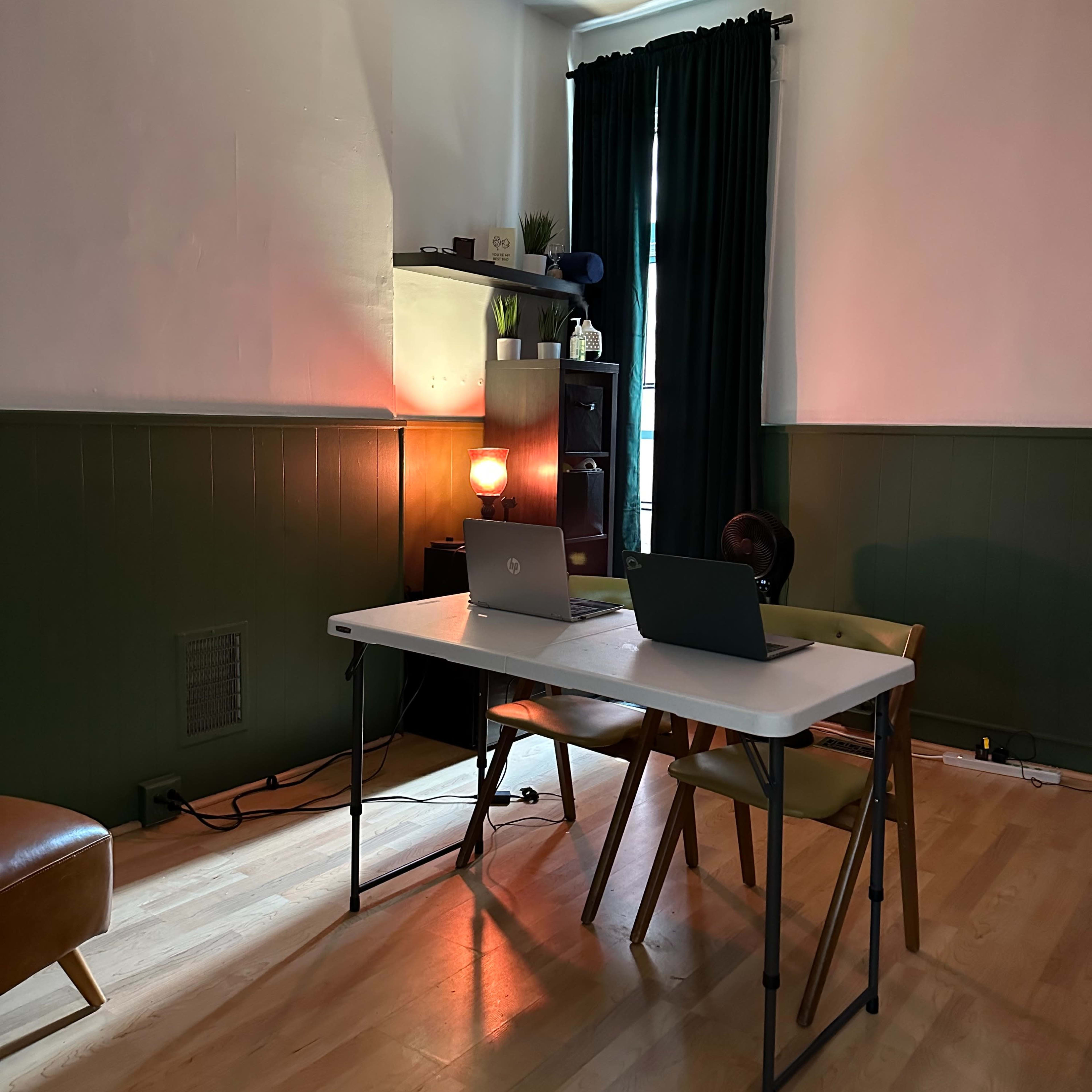 A simple workspace is set up in a room with a table, two laptops, a chair, and dark green walls.