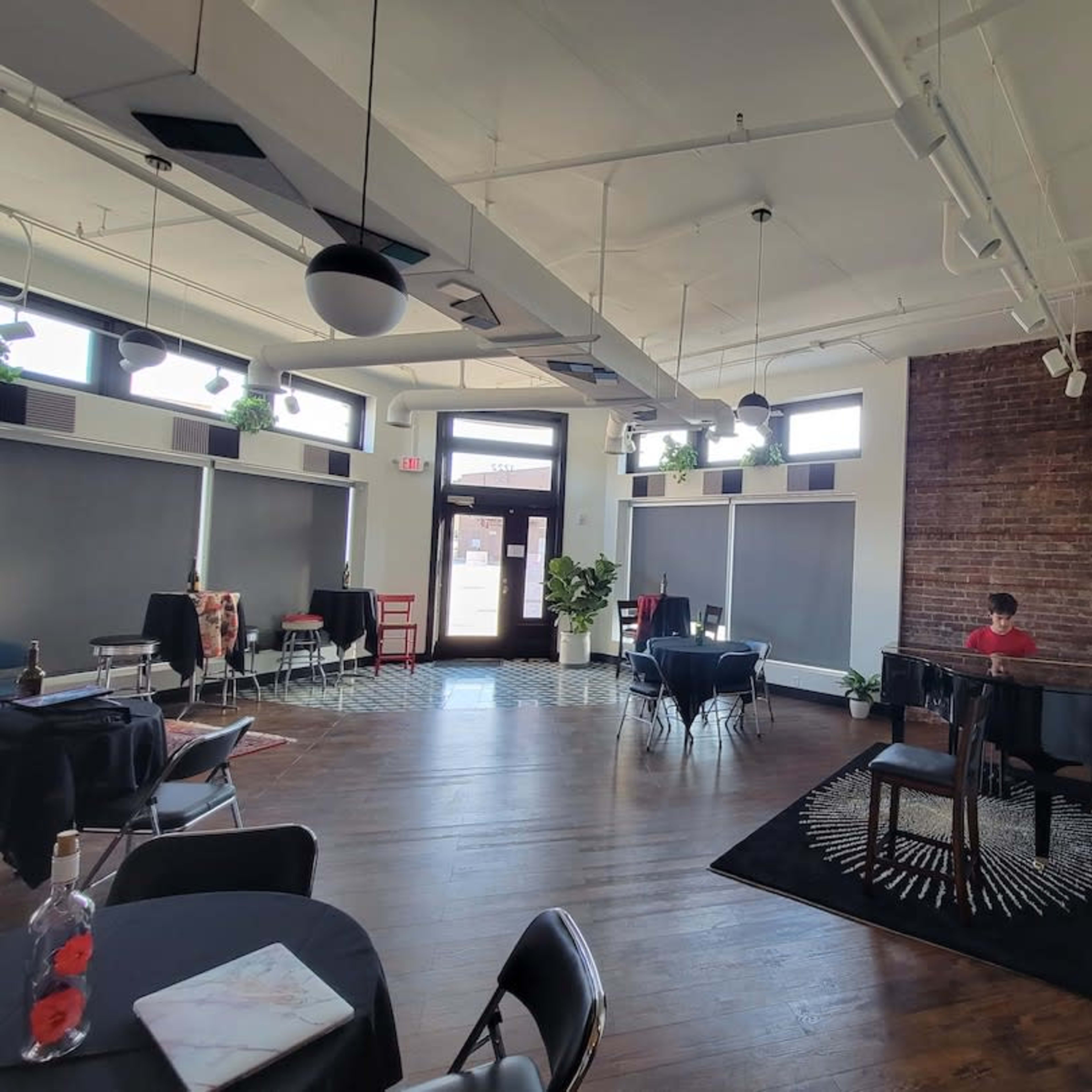 A spacious interior features wooden floors, tables set for dining, and a person seated at a piano against a backdrop of exposed brick.