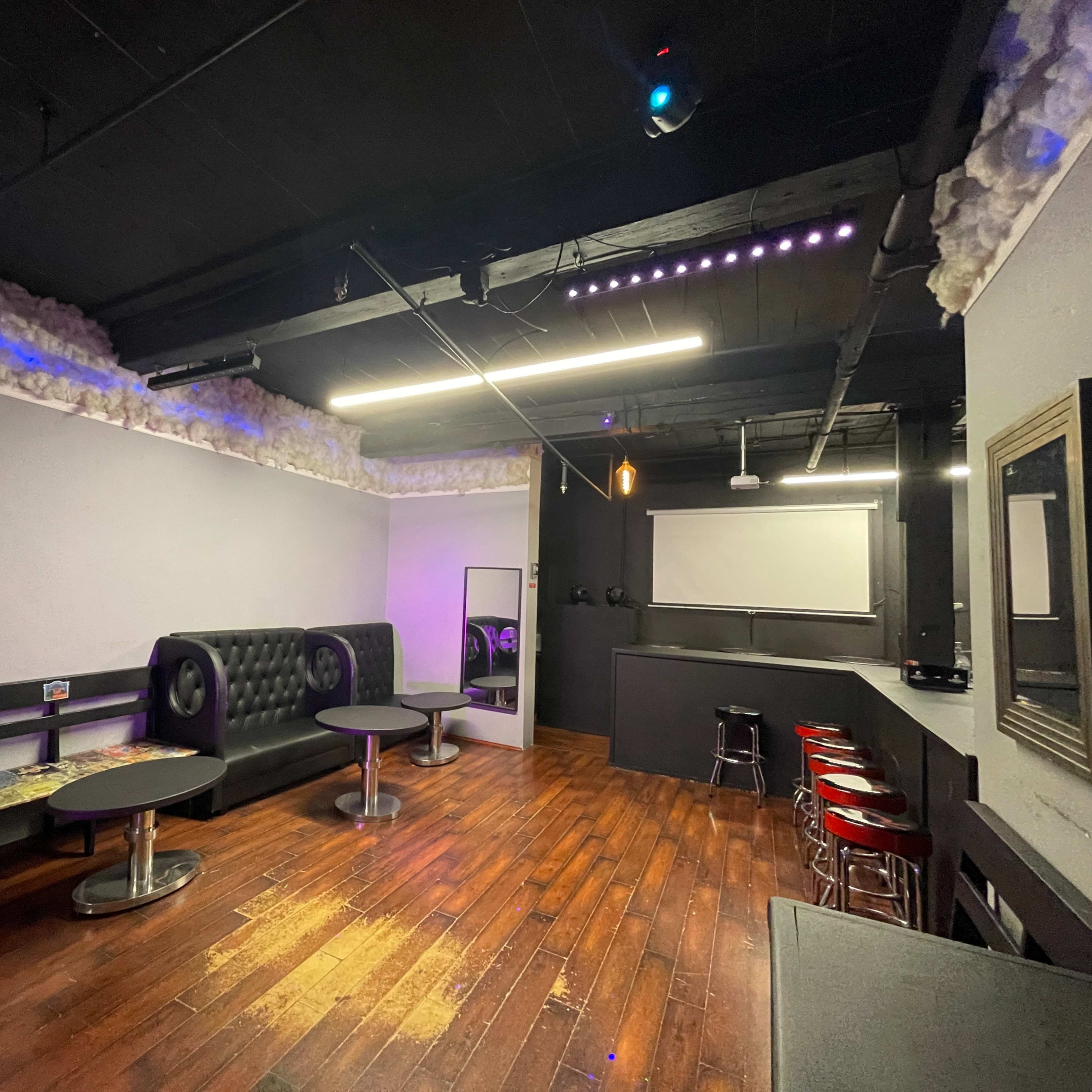 A dimly lit lounge area with black leather furniture, wooden flooring, and a projection screen on the wall.