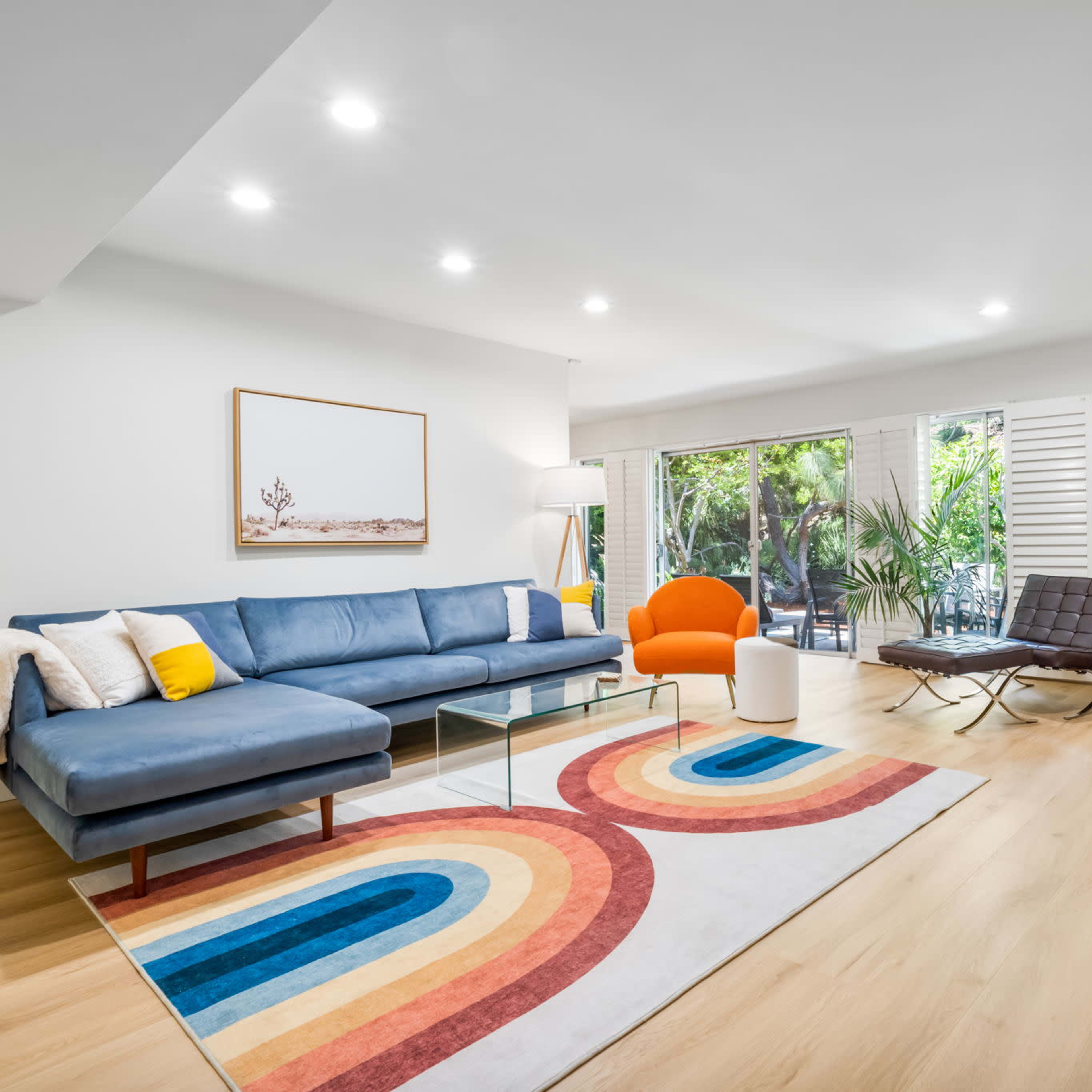 A modern living room with a blue sectional sofa, an orange accent chair, a glass coffee table, and a multicolored area rug, featuring large windows that overlook a green outdoor space.