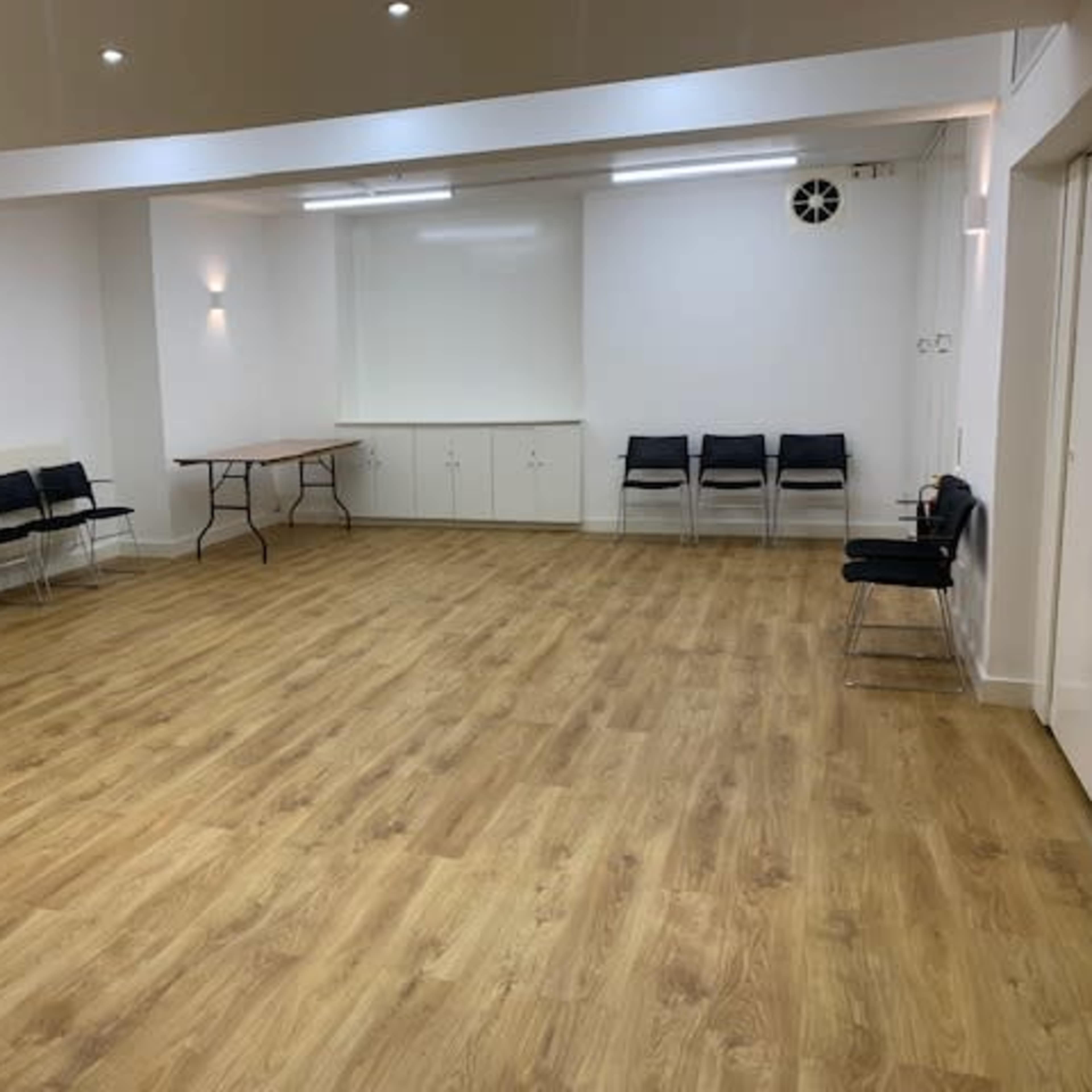 An empty room with wooden flooring, white walls, a table, and several black chairs lined against one side.