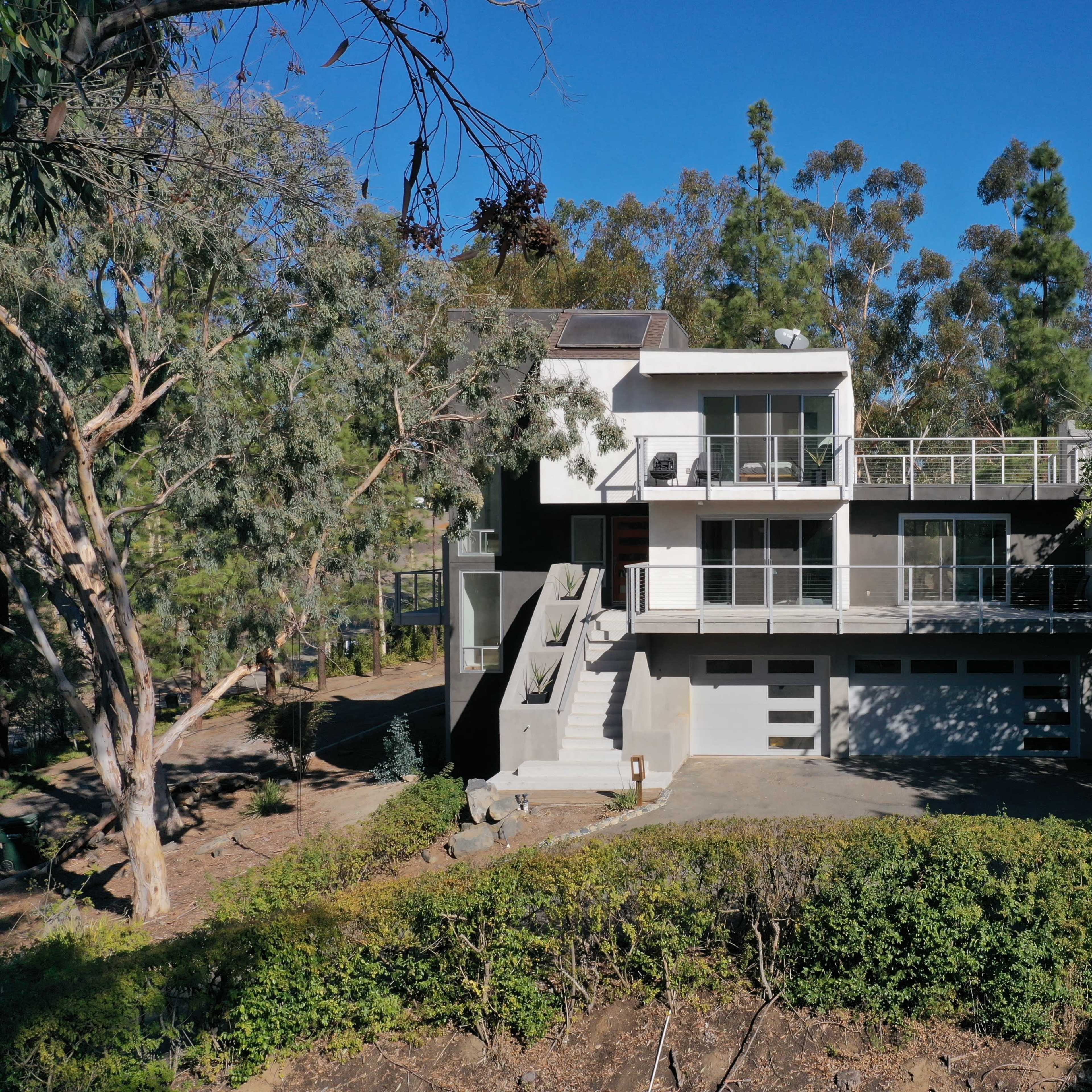 A modern two-story house with a flat roof is situated amidst trees, featuring a large front terrace and a driveway leading to a garage.