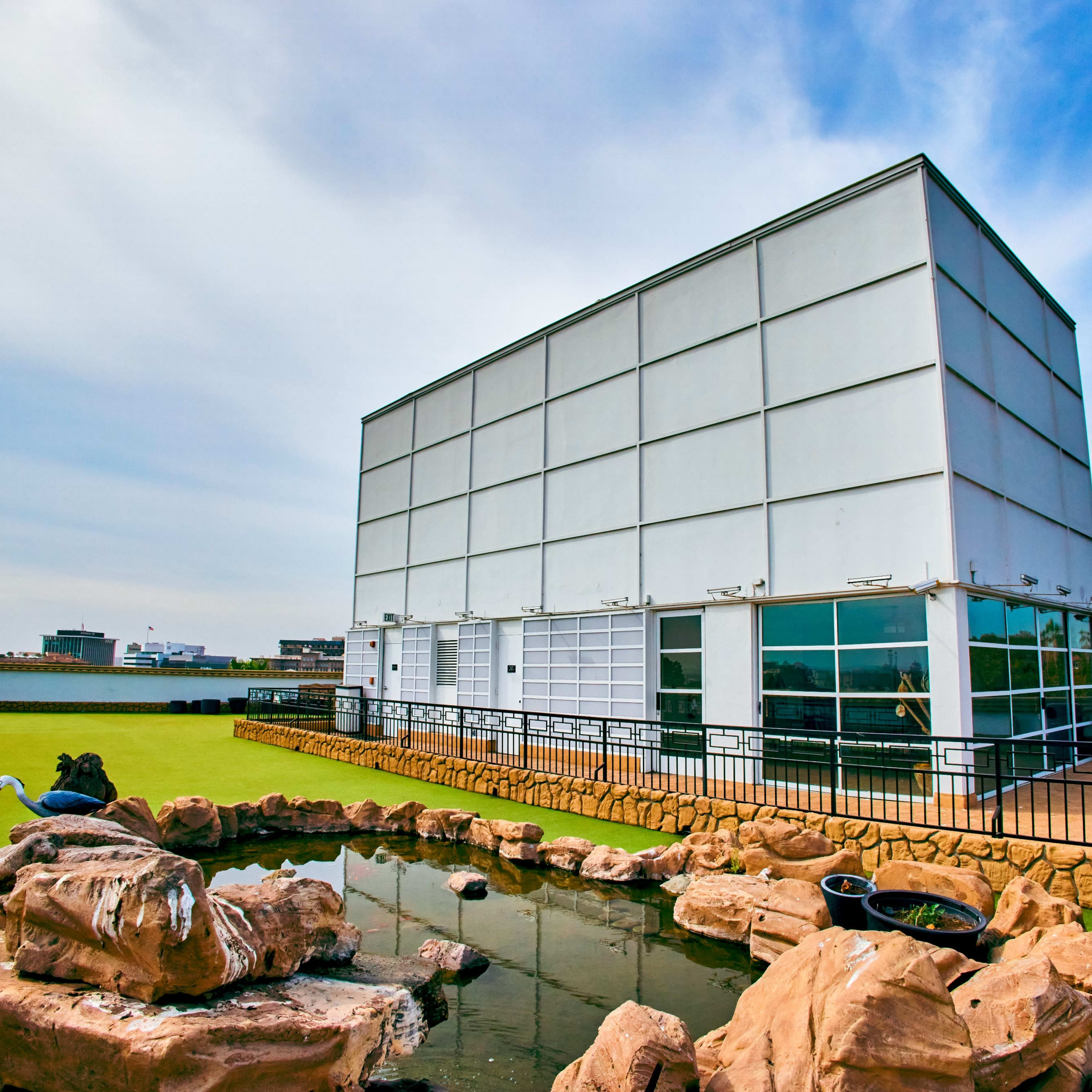 A modern white building with large windows is situated on a rooftop featuring a small, rocky pond and green landscaping.