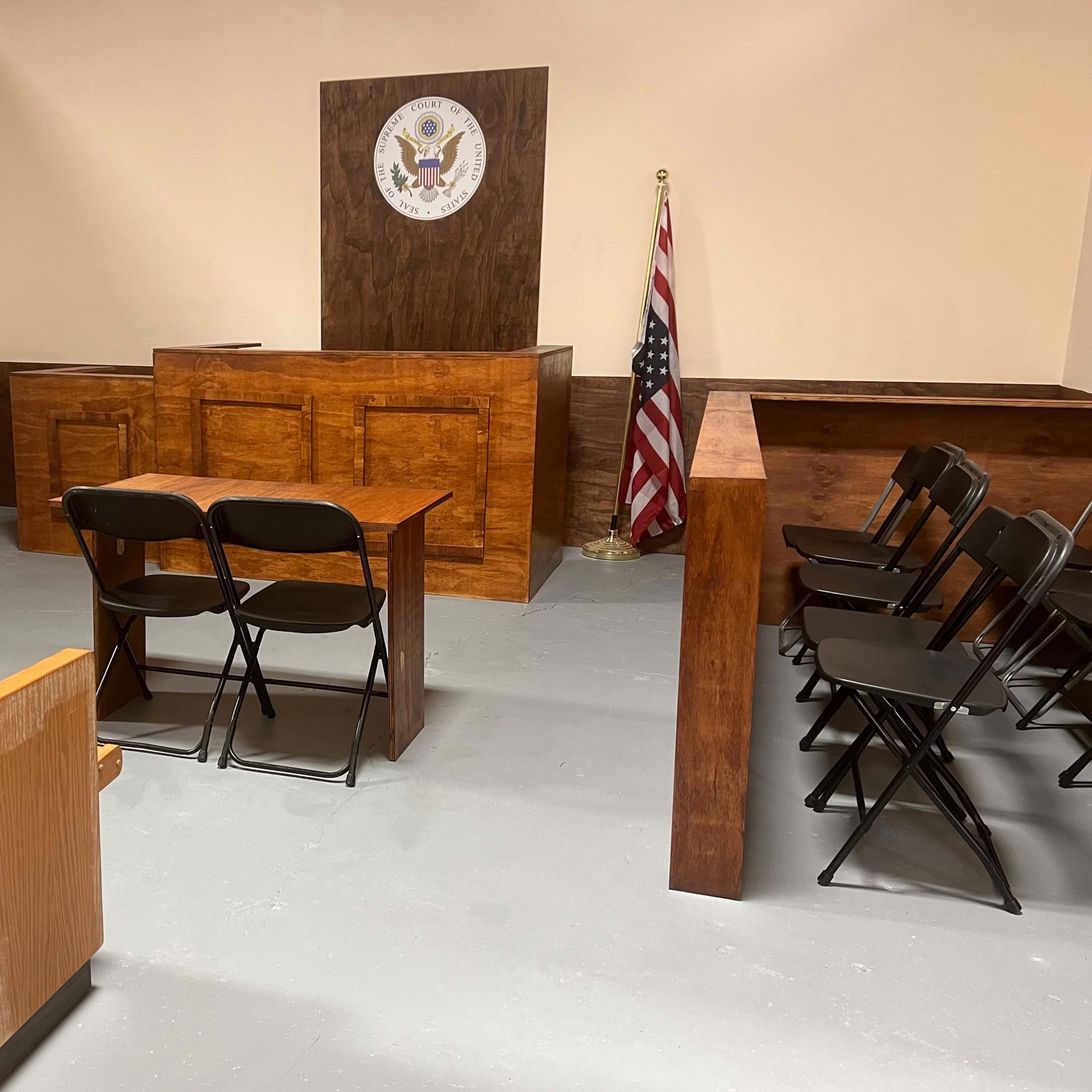 A courtroom with two wooden tables, black folding chairs, a United States flag, and a seal of the U.S. on the wall behind the judge's bench.