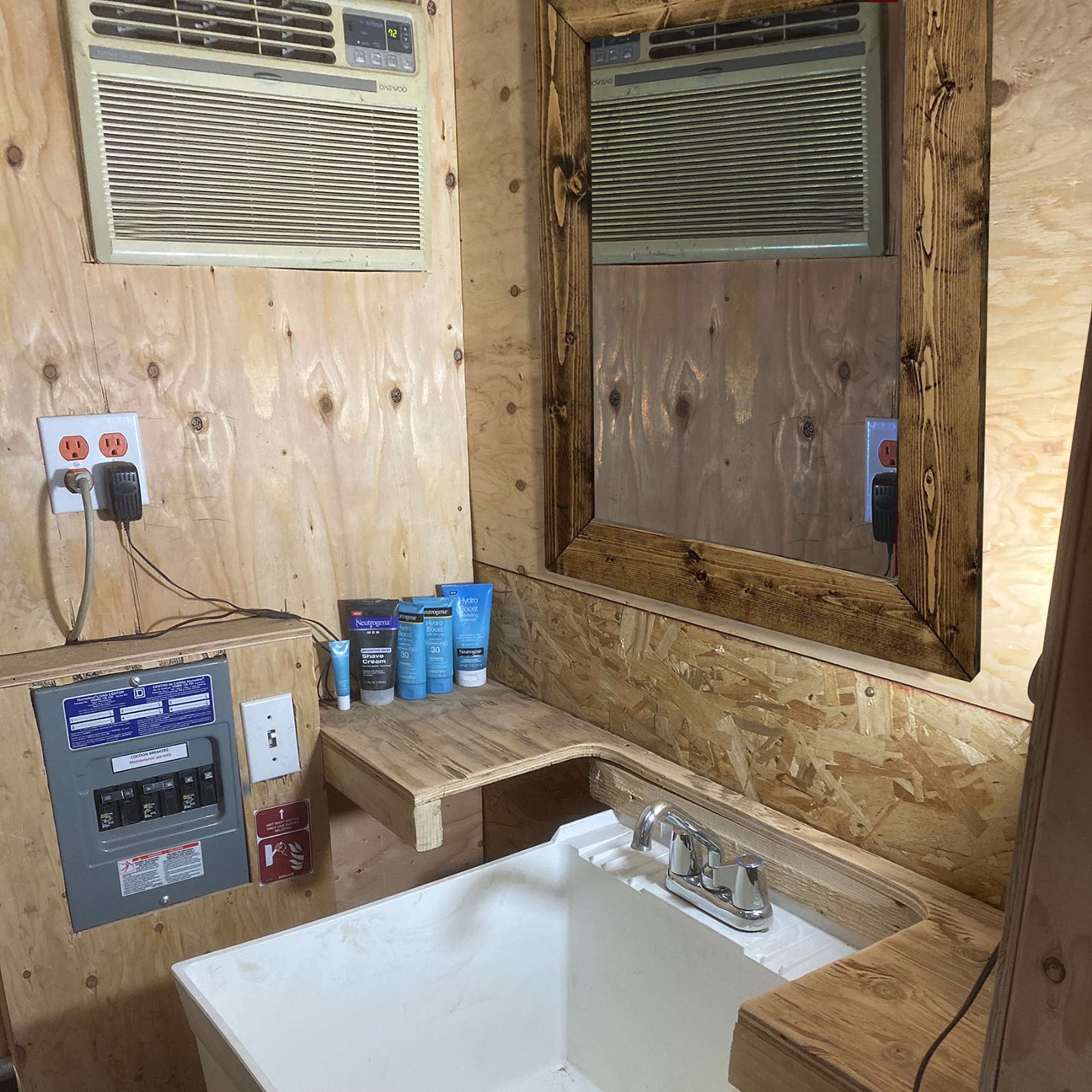 A small bathroom with a sink, a wooden mirror frame, an air conditioning unit above, and various personal care products on a shelf.
