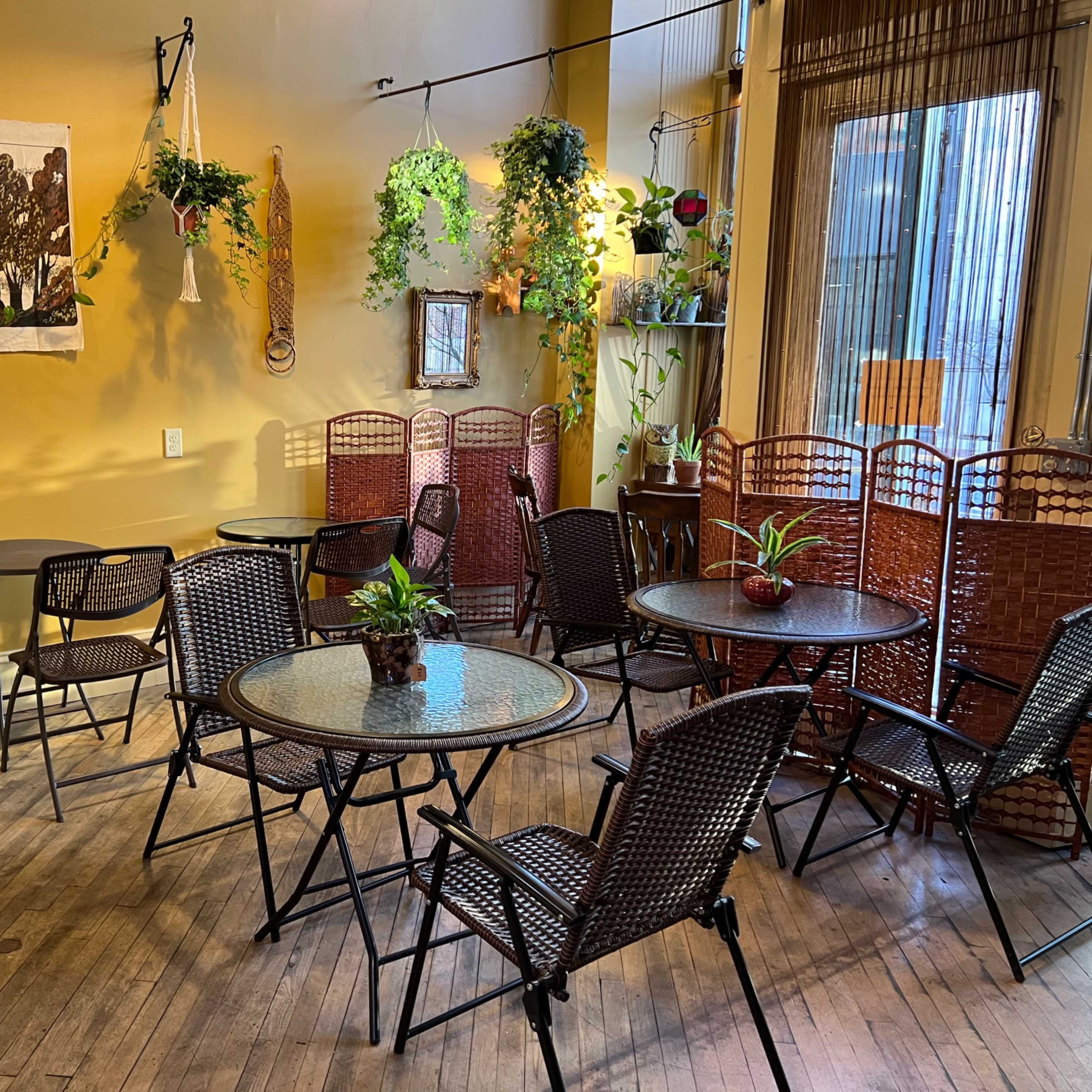 A cozy café space with several round tables and chairs, adorned with plants and a decorative wall hanging.