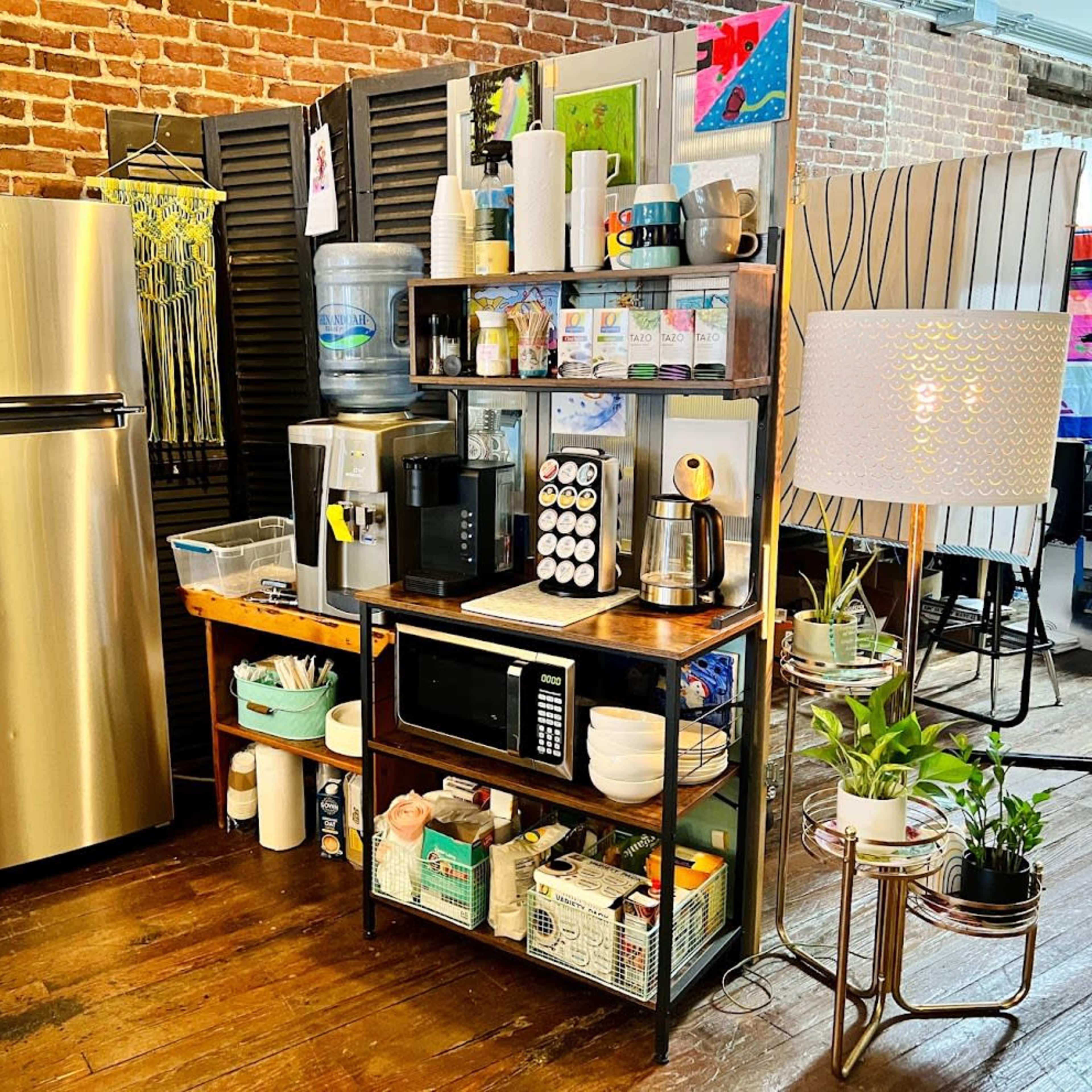 A kitchen area with a stainless steel refrigerator, a shelving unit filled with various kitchen supplies, a microwave, and a coffee maker, set against a backdrop of exposed brick walls.