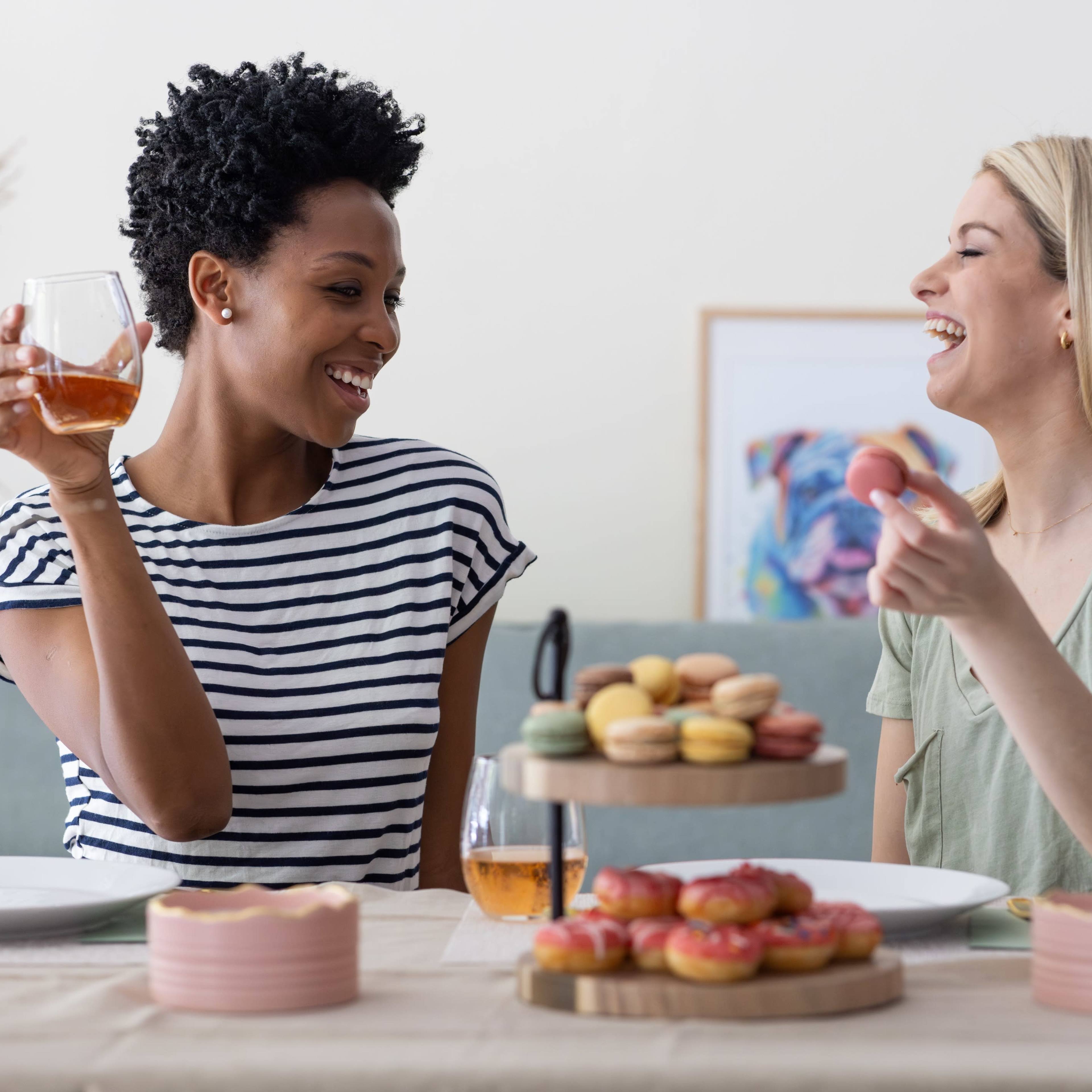 Two women sit at a table filled with desserts, smiling and enjoying drinks while engaged in conversation.