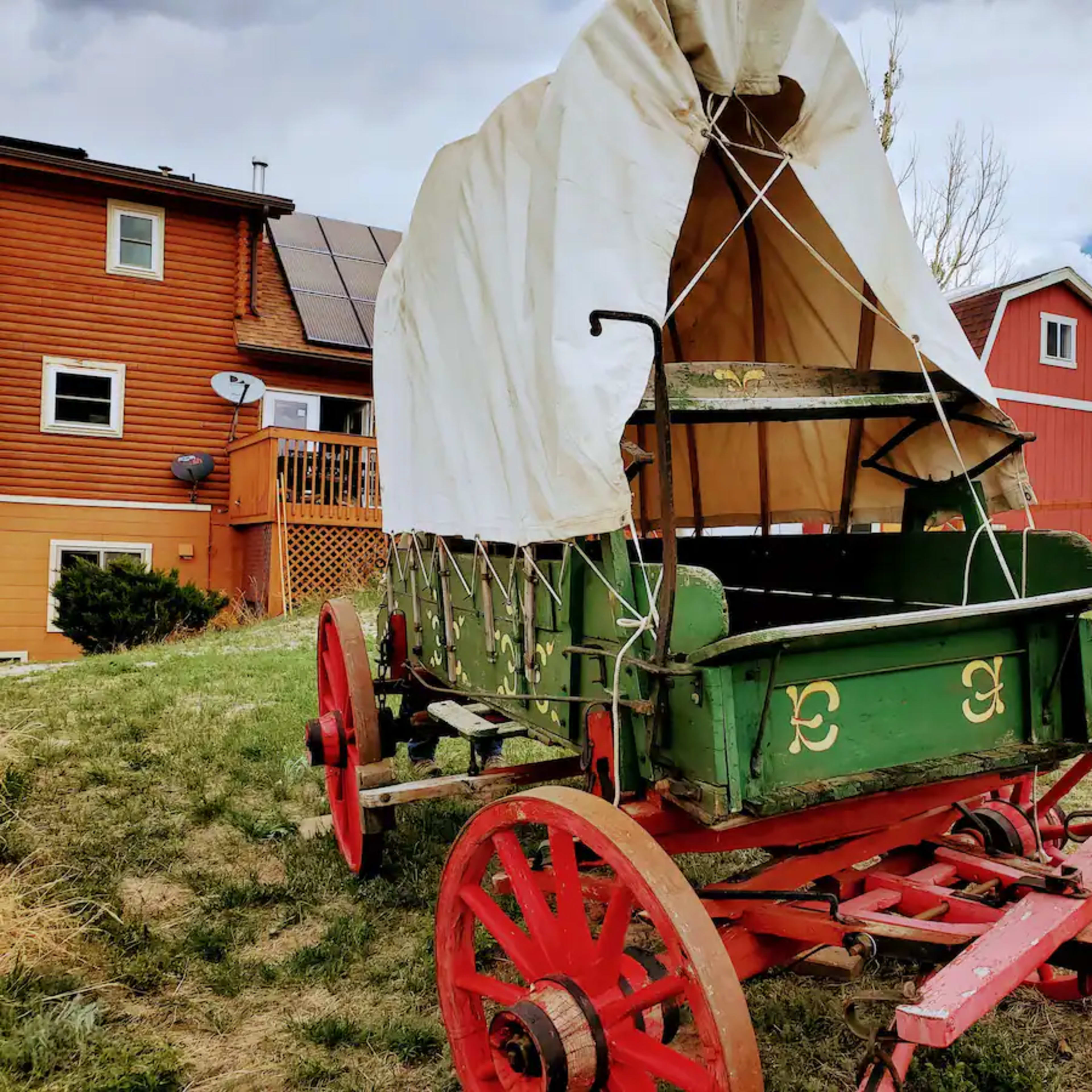A covered wooden wagon with red wheels is positioned on grass near a house and barn in a rural setting.