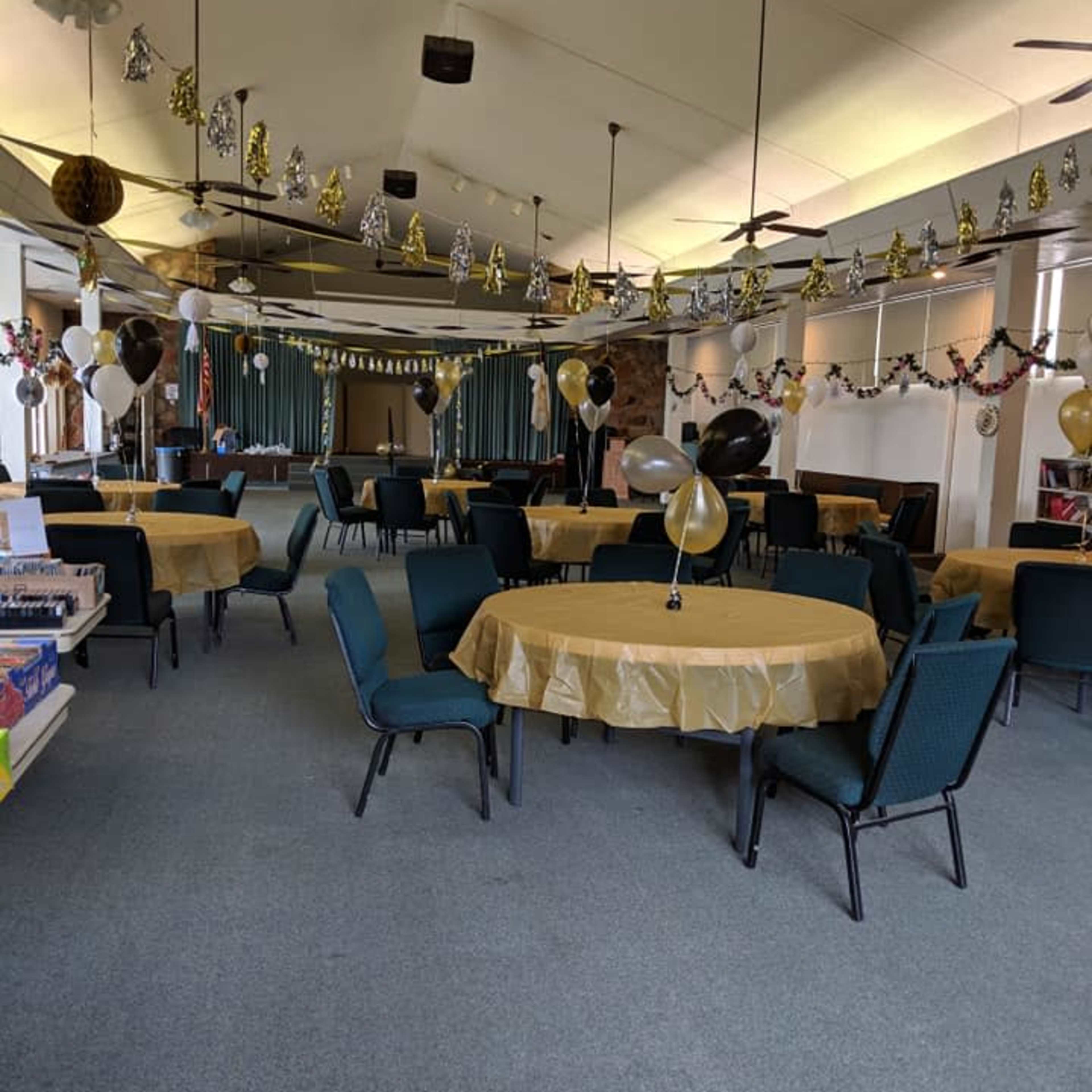 A banquet hall set up with round tables covered in gold tablecloths, adorned with black and gold balloons, streamers, and decorative elements for a celebration.